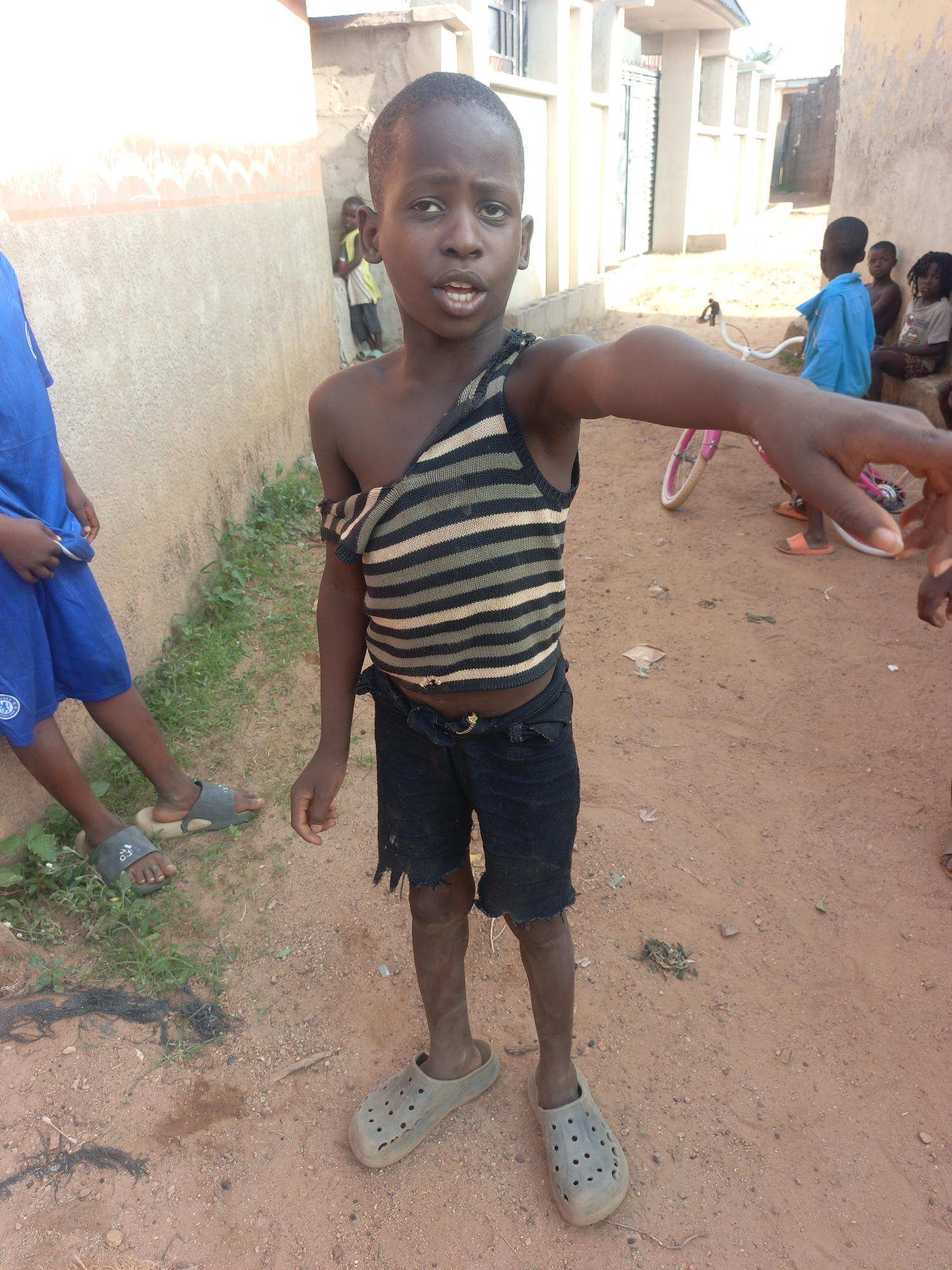 The image shows a boy standing on a dirt road in Akwanga, Nigeria. He is wearing tattered black shorts, a striped tank top with one shoulder exposed, and gray Crocs. He is pointing his right arm out towards the viewer. Behind him, there are several other children. One child is wearing a blue shirt, and another is standing near a wall. There is a pink bicycle in the background. The area appears to be a residential street with simple buildings. Vegetation is sparsely visible along the wall on the left. The ground is primarily red dirt.