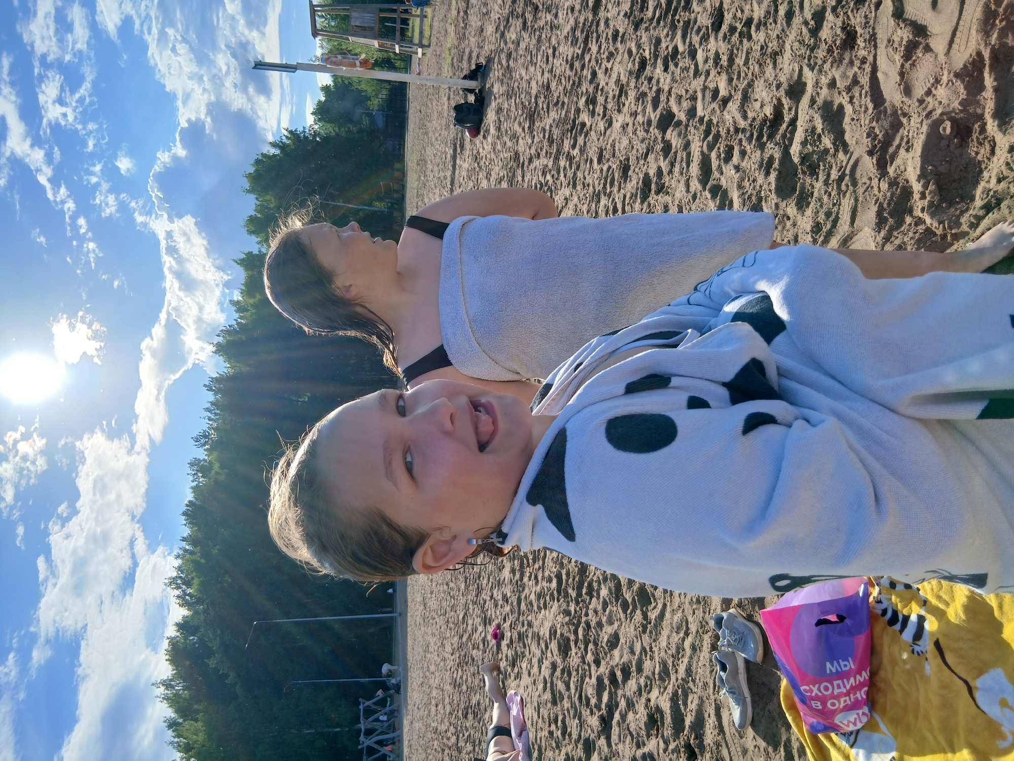 The media file shows two young females at a sandy beach on a sunny day. The person in the foreground is a young girl with light brown hair, wearing a white towel with black polka dots. She is sticking out her tongue and looking at the camera, appearing playful. Behind her, another female, likely an adult, is wrapped in a white towel and looking away from the camera. Both individuals seem to be enjoying a day at the beach.
The setting is a beach with sand and some vegetation in the background. The sky is blue with scattered white clouds, and the sun is visible, casting a warm glow and lens flare. In the background, there are glimpses of playground equipment and trees, suggesting a park or recreational area near the beach. There is also a yellow towel with a black and white striped pattern and a pink plastic bag with Russian text visible on it. The text on the bag reads: "МЫ СХОДИМ В ОДНО".
The overall mood of the image is casual and cheerful, indicative of a summer day spent outdoors. The time of day appears to be late afternoon, given the angle of the sun and the warm light. The location cues, such as the vegetation and the architecture in the far background, along with the Russian text on the bag, suggest the location is Chaikovsky, Russia.