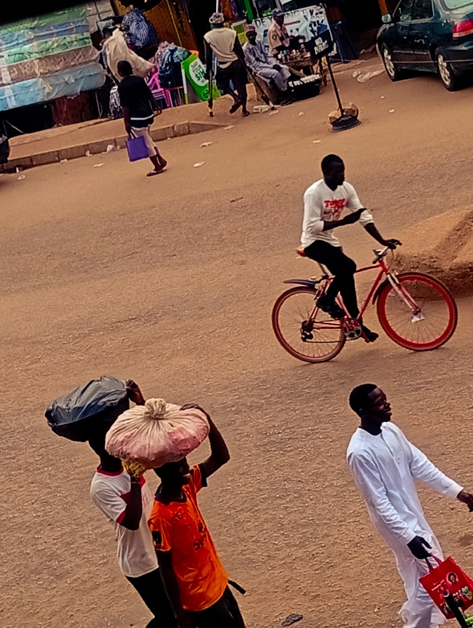 The image shows a street scene in Bukuru, Nigeria. The road is a reddish-brown color and appears to be unpaved in sections. Several people are visible. One man is riding a red bicycle with a white t-shirt and black pants. Three individuals are walking towards the viewer: two people carrying bundles on their heads, and one man in a white robe carrying a red bag. In the background, there are a few more people, including some gathered near a small roadside vendor stall and a dark-colored car parked on the side of the road. There are also some mattresses stacked near the edge of the road.