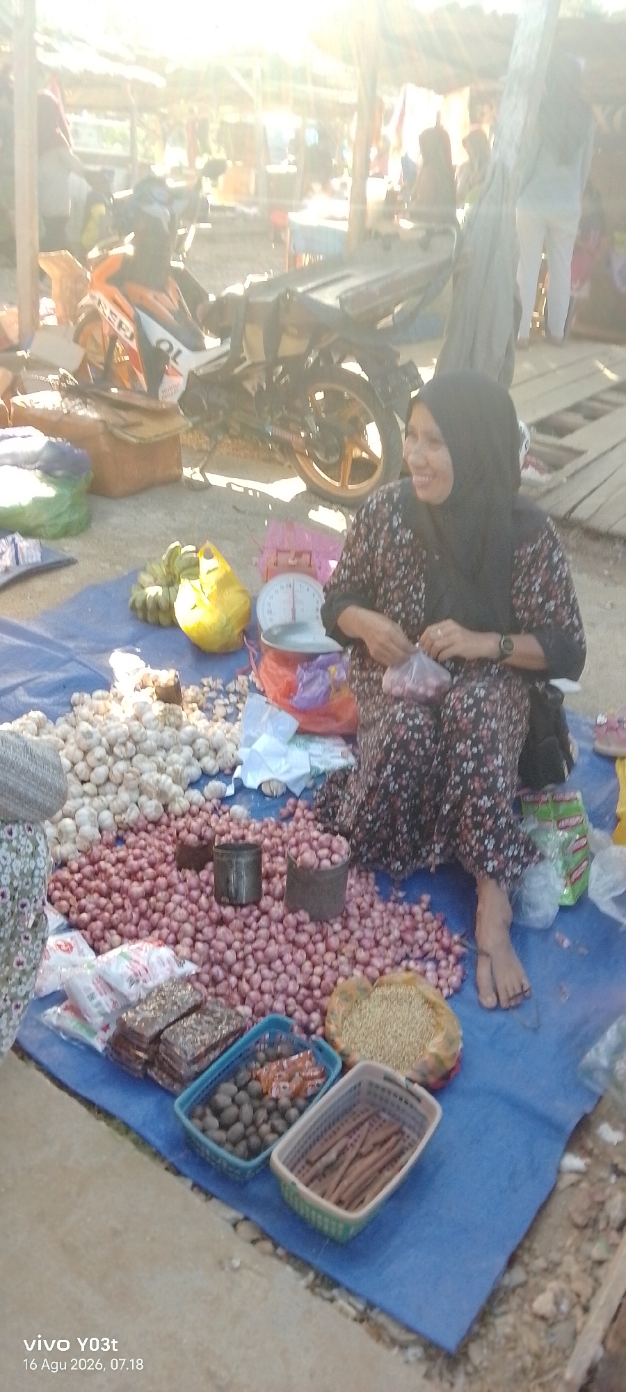 In Palosilae, Indonesia, a woman in a floral-patterned dress and black hijab sits on a blue tarp at a market. She is smiling and holding a small plastic bag filled with red onions. In front of her, the tarp is covered with a large pile of red onions, as well as a significant amount of garlic. To the left, a bunch of bananas and a yellow plastic bag are visible. A silver weighing scale rests on an orange bag. Behind the woman, a motorcycle with orange and white accents is parked. Several other people are in the background, as well as wooden stalls and structures that suggest an outdoor market setting. The scene is illuminated by bright sunlight, casting lens flares across the image.