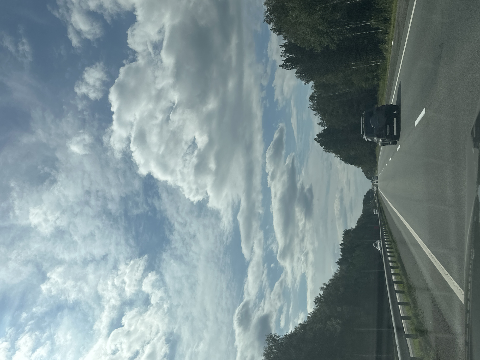 The image shows a view from inside a vehicle traveling on a highway. The road is paved and has white lane markings. There are trees lining both sides of the highway. The sky is partly cloudy with large, white clouds against a blue sky. Several other vehicles are visible on the road, including a black SUV in the foreground. The scene appears to be a typical daytime highway commute in a rural or semi-rural area. There are no people visible outside of the vehicles.
