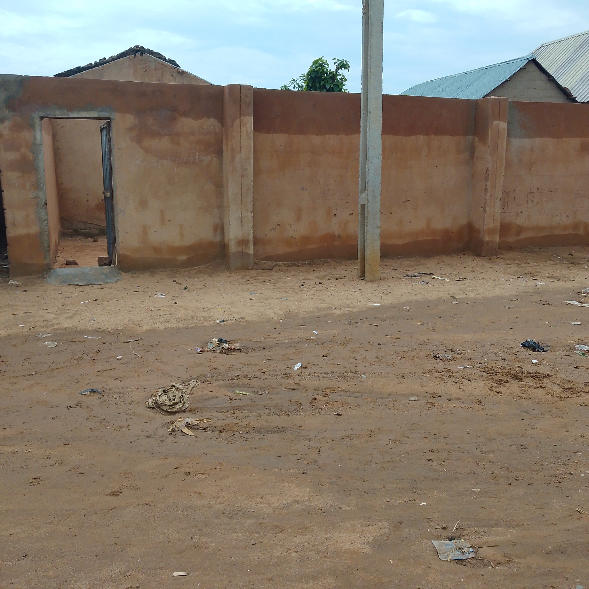 The image depicts an outdoor scene on a dirt road in Pantame, Nigeria. A weathered, light brown plastered wall dominates the mid-ground, with a partially visible open doorway on the left. The wall is punctuated by vertical support pillars. Beyond the wall, the roofs of two buildings are visible. A concrete utility pole stands in front of the wall. The ground is covered in sand and strewn with various pieces of debris, including plastic bags, wrappers, and what appears to be a bundle of fabric or clothing. The sky above is overcast with clouds. The overall impression is of an unkempt, possibly underdeveloped area.