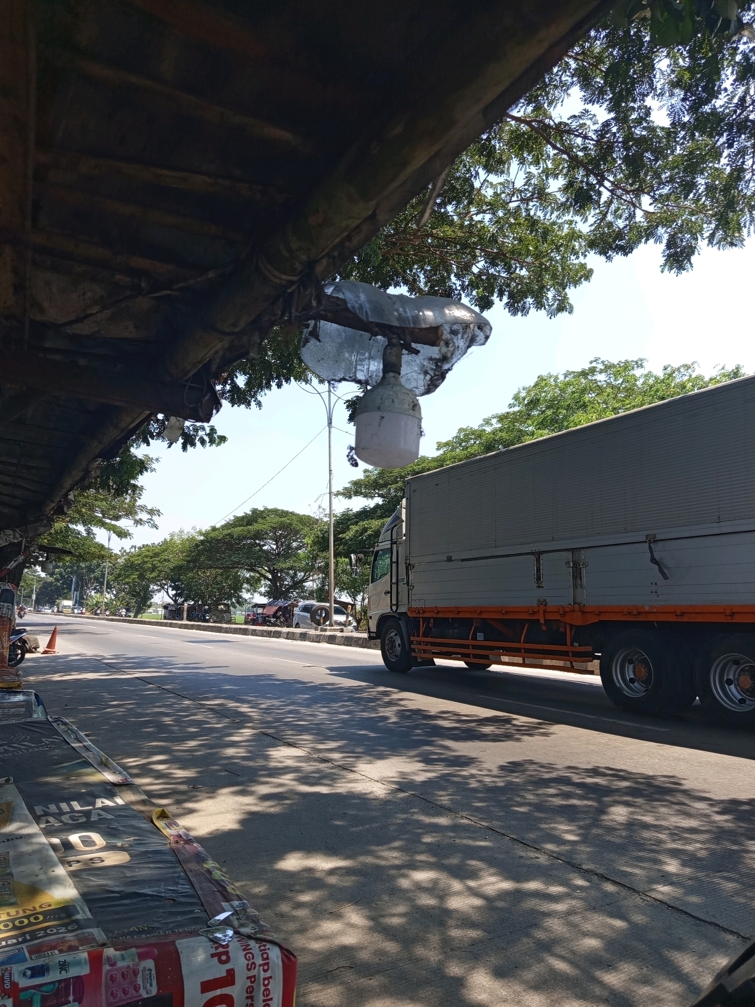 A truck is driving on a road in Karawang, Indonesia. The truck is gray and orange and has six wheels. The road is asphalt and has a white line down the middle. There are trees on the side of the road, and the sky is blue. The image is taken from the perspective of someone standing on the side of the road, under a canopy. There are also some posters on the ground in front of the viewer.