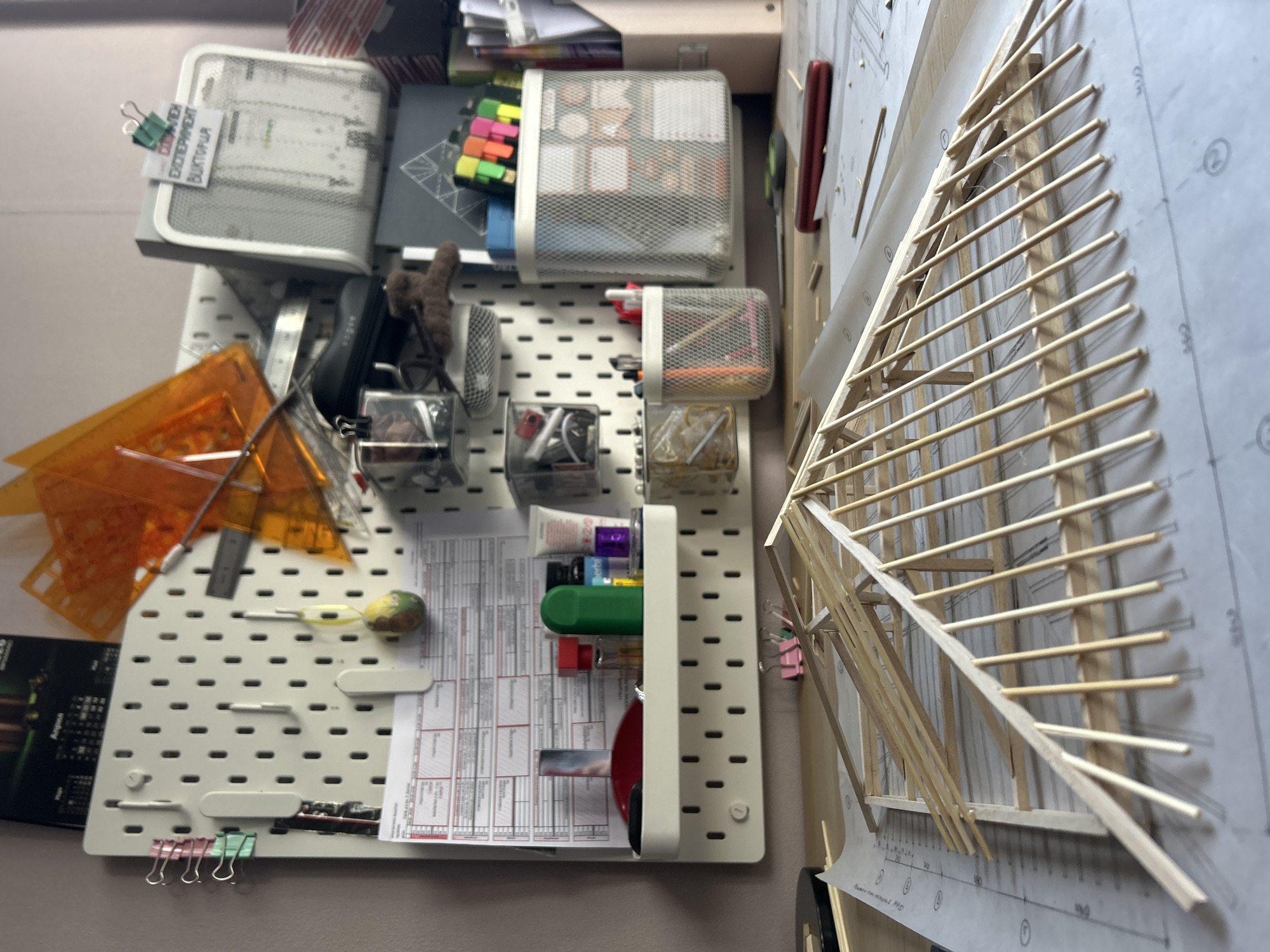 This image captures an overhead view of a well-organized workspace in Sofia, Bulgaria, featuring a pegboard organizer on the left and an architectural model on the right, resting on blueprints.
The left side of the frame is dominated by a light-colored pegboard, vertically mounted and accessorized with various containers and tools. Several mesh metal containers hold office supplies: the top left contains papers with a small note clipped to it, while the top right and middle containers are filled with colorful sticky notes, pens, and markers. Smaller mesh containers below hold items like earphones and other desk accessories. White plastic shelves on the pegboard hold small bottles or tubes, possibly art supplies, and a red circular item. Directly attached to the pegboard are transparent orange plastic rulers, a metal ruler, a small craft knife, a yellow rubber band, and small binder clips in pink and green. A printed document with tables and Cyrillic text is clipped to a lower shelf.
On the right side of the frame, a delicate wooden model, likely an architectural scale model of a roof or building frame, is laid out on a large sheet of paper. This paper appears to be an architectural blueprint or technical drawing, featuring grid lines, numbers (e.g., "0", "100", "200", "300", "500"), and some Cyrillic text. The wooden structure consists of numerous thin wooden sticks or dowels, meticulously arranged to form a skeletal framework. A red cylindrical object is partially visible behind the model.
Visible text includes "ЭКСПЕРИМЕНТ ВИКТОРИЯ" ("EXPERIMENT VICTORIA") on a small paper clipped to a container on the pegboard. On the blueprint, numbers like "500", "2м", "1м", and "мащаб 1:100" ("scale 1:100") are discernible. The scene suggests a creative or technical project in progress, indicating a detail-oriented environment.
