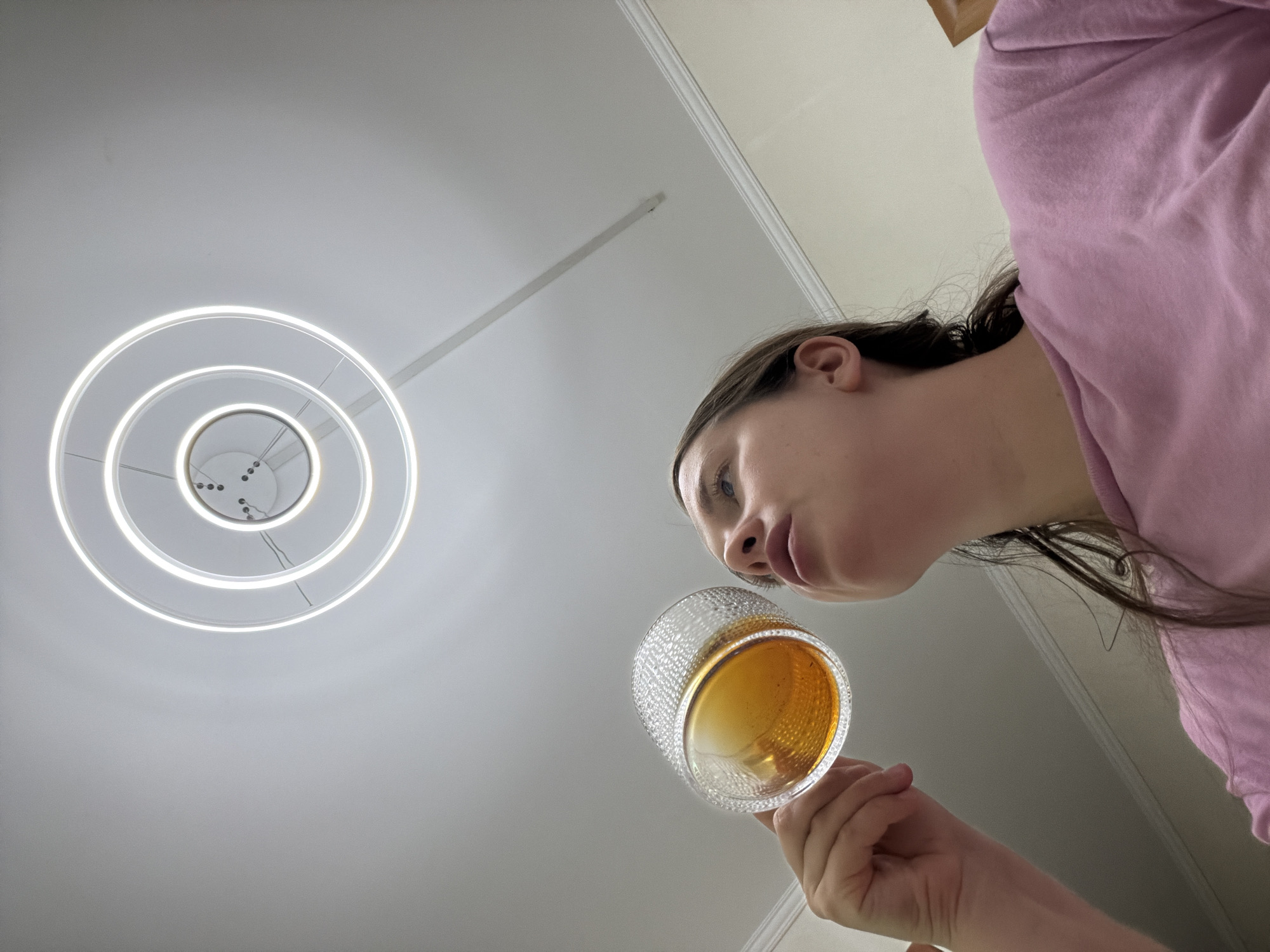 A young female, likely a teenager or young adult, is depicted from a low-angle perspective, looking upwards. She occupies the right side of the frame, wearing a pink long-sleeved top. Her dark hair is pulled back, and her face is shown in profile as she gazes towards the left. She holds a clear, textured glass containing a yellowish-brown liquid, possibly tea or another beverage, close to her mouth or nose, suggesting she is about to drink or smell its contents.
The background primarily consists of a plain, light-colored ceiling. A prominent modern light fixture, composed of concentric illuminated circles, is affixed to the ceiling on the left side of the image, emitting a bright white glow. A subtle ceiling molding detail is visible in the upper right portion of the frame.
The photograph is taken from a unique, extreme upward angle, creating a dynamic composition. There are no specific cues within the image itself to confirm the location in Sochi, Russia, but the setting appears to be an indoor space. The lighting is artificial, coming from the circular fixture, and there is no visible text or strong emotional expression from the subject.