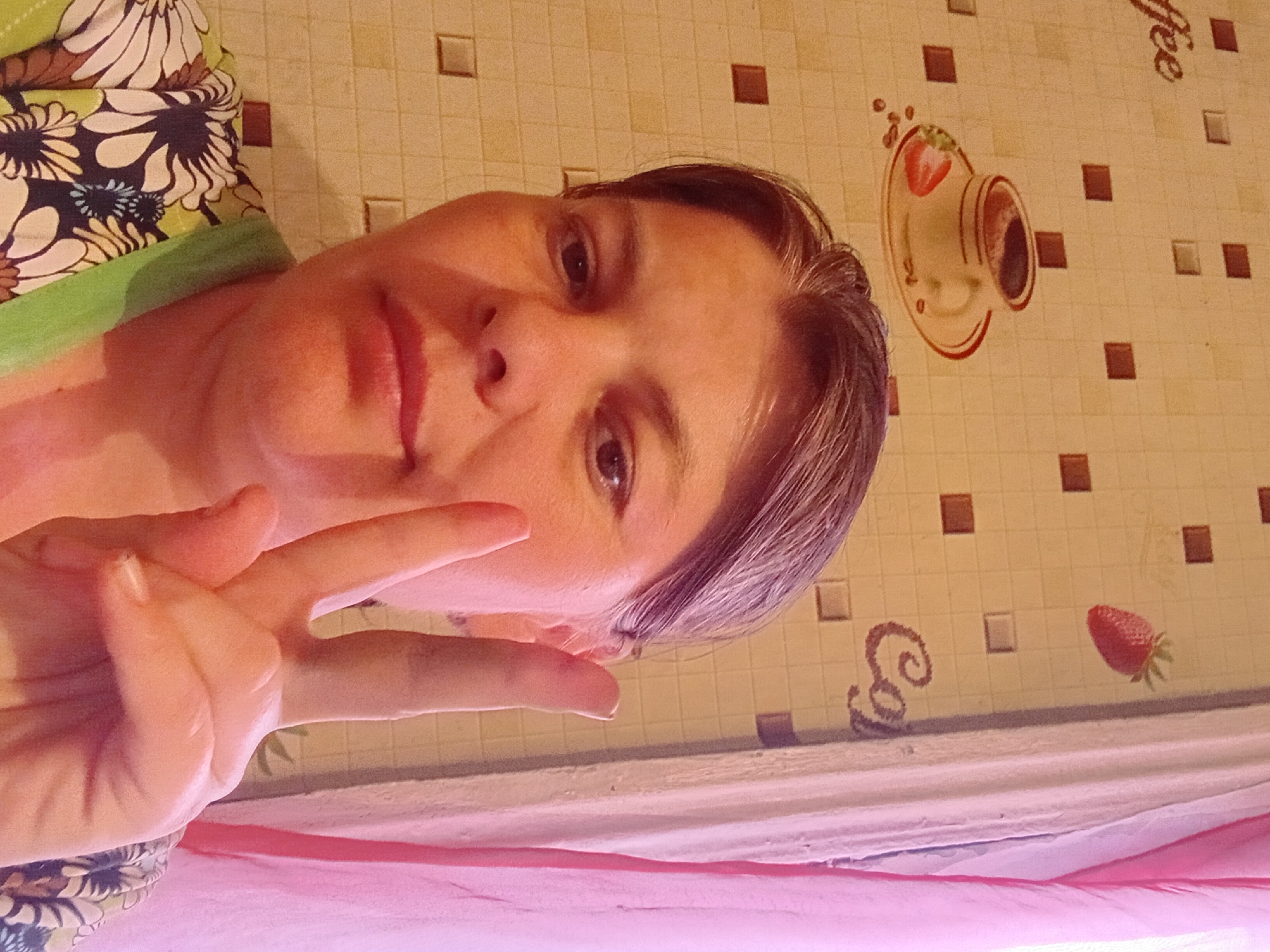 The image is a close-up selfie of a woman with brown hair and fair skin. She is making a peace sign with her right hand in front of her face. She is wearing a green and floral-patterned top. The background appears to be a tiled wall with images of a coffee cup, a strawberry, and the word "Coffee" and "Co" on it. A pink curtain is visible on the right side of the image. The image is taken indoors, likely in a residential setting. There is no information available to determine the location in the city of Малая Владимировка ауылдық округі, Kazakhstan.