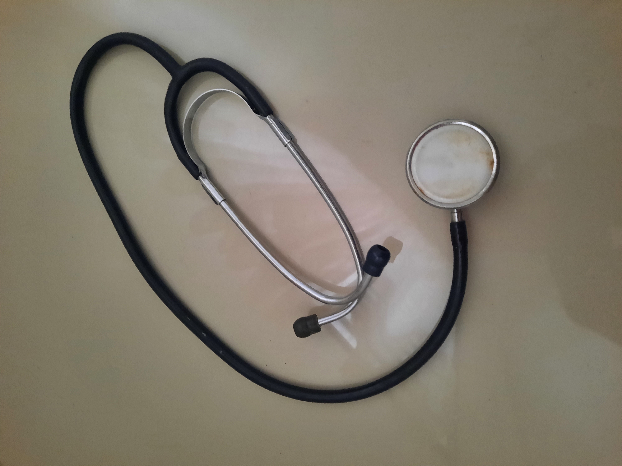 A black stethoscope is laid out on a light brown, possibly tiled surface. The stethoscope consists of a flexible black tube connecting a chestpiece to eartubes. The chestpiece, a circular diaphragm enclosed in a silver metal rim, is positioned at the bottom right of the image. The eartubes are made of silver metal and terminate in black ear tips, which are angled to fit into the ears. The stethoscope is positioned diagonally across the frame. The surface appears flat and slightly reflective, with some subtle shading and minor imperfections, such as small dark specks, visible on its surface. The lighting suggests a well-lit indoor environment. The image does not contain any people or indications of a specific location within Lhokseumawe, Indonesia.