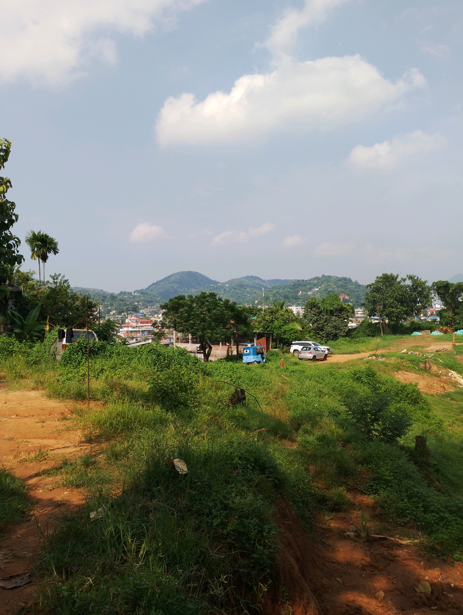 The image depicts a landscape with a town nestled in a valley, surrounded by lush green hills under a partly cloudy sky. In the foreground, a dirt path winds through overgrown grass and foliage. To the left, a tall tree with broad leaves stands prominently, and a couple of palm trees are visible. Further into the scene, on a dirt road, are two parked cars and a small blue three-wheeled vehicle. The town in the valley features a cluster of buildings with various colored roofs, interspersed with trees. The background is dominated by gently sloping, verdant hills. The overall impression is one of a semi-urban or rural setting within a hilly region, likely in or around Dispur, India, given the vegetation and architectural style.