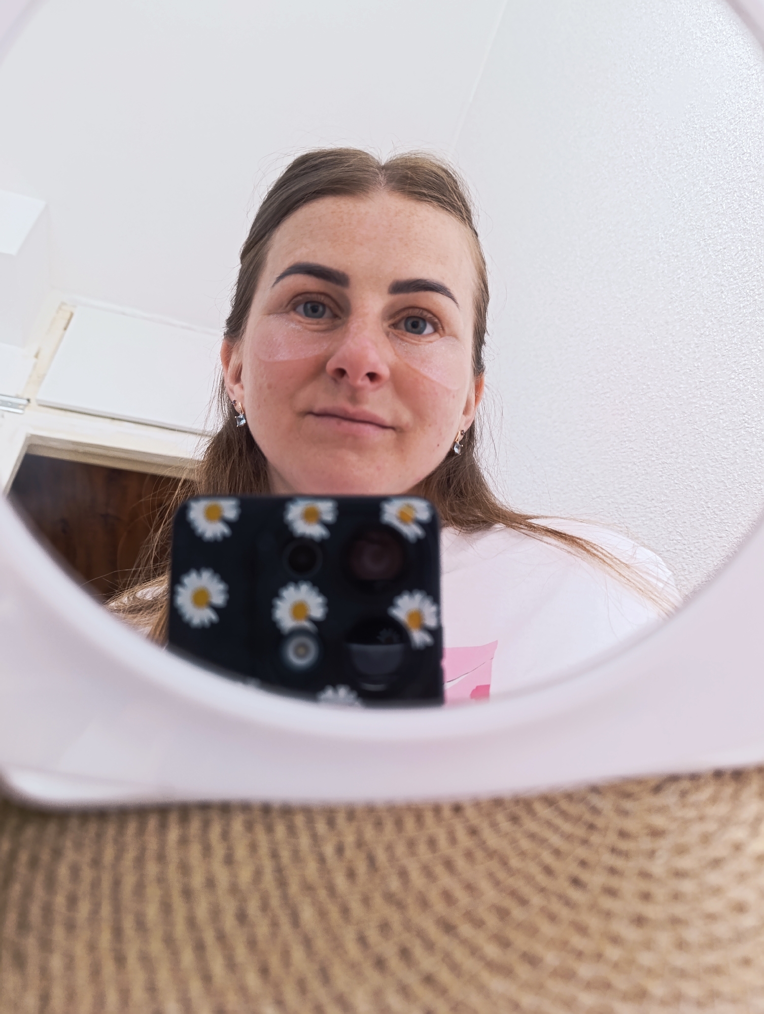 Here's a matter-of-fact description of the image:
A woman with fair skin and freckles is taking a selfie in a round, white mirror. She has clear eye patches under her eyes. She's wearing earrings and has her hair pulled back. The phone she's using to take the photo has a black case with a daisy pattern. The background visible in the mirror includes a white wall and a glimpse of a door. The surface the mirror is resting on appears to be woven.
The city and specific location are unknown.