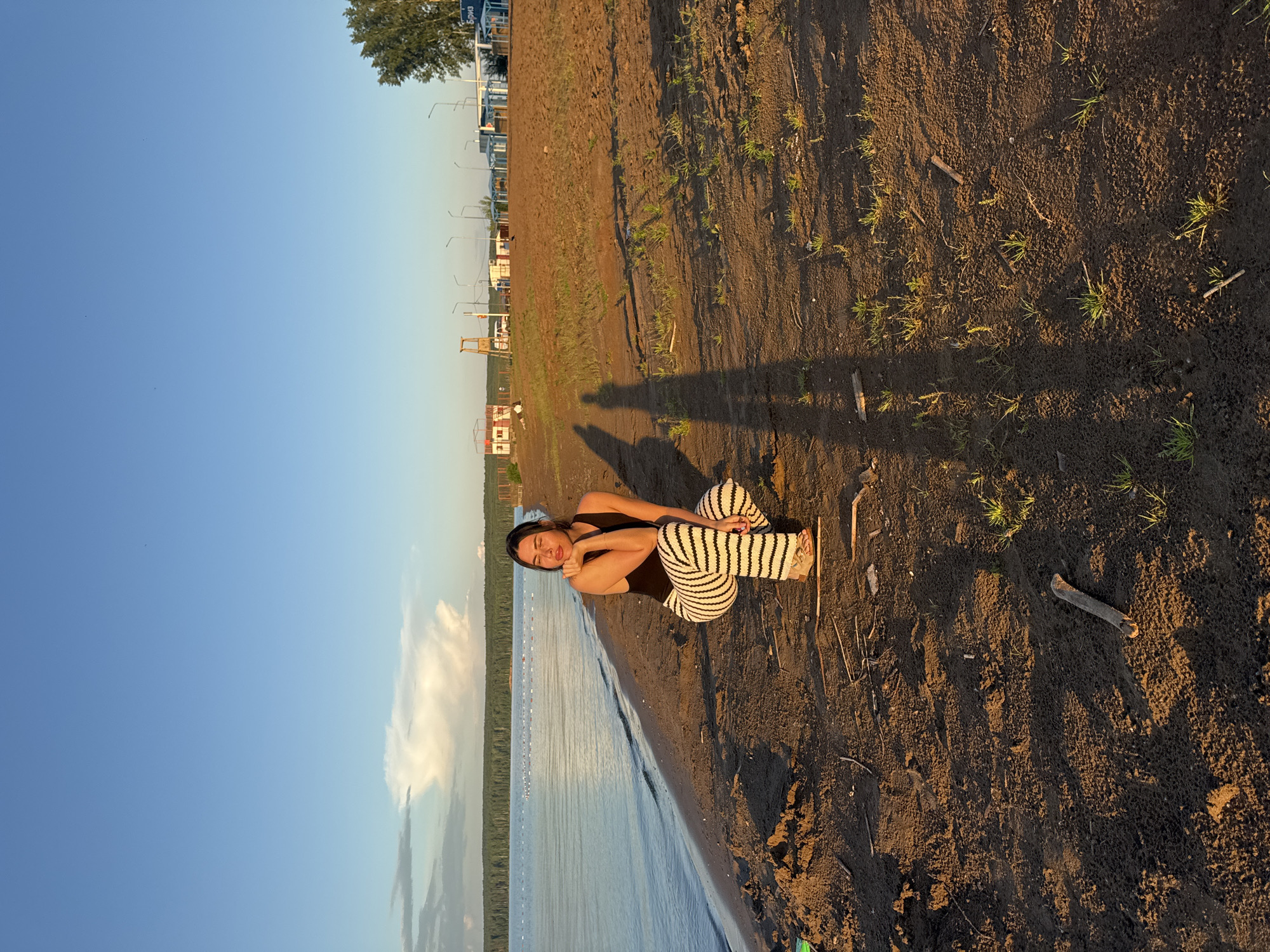 A woman is crouching on a sandy beach by a body of water. The sun is low in the sky, casting long shadows. In the background, there are some buildings and trees. The overall impression is one of peace and tranquility. The location is likely on the Kama River in Chaikovsky, Russia.
