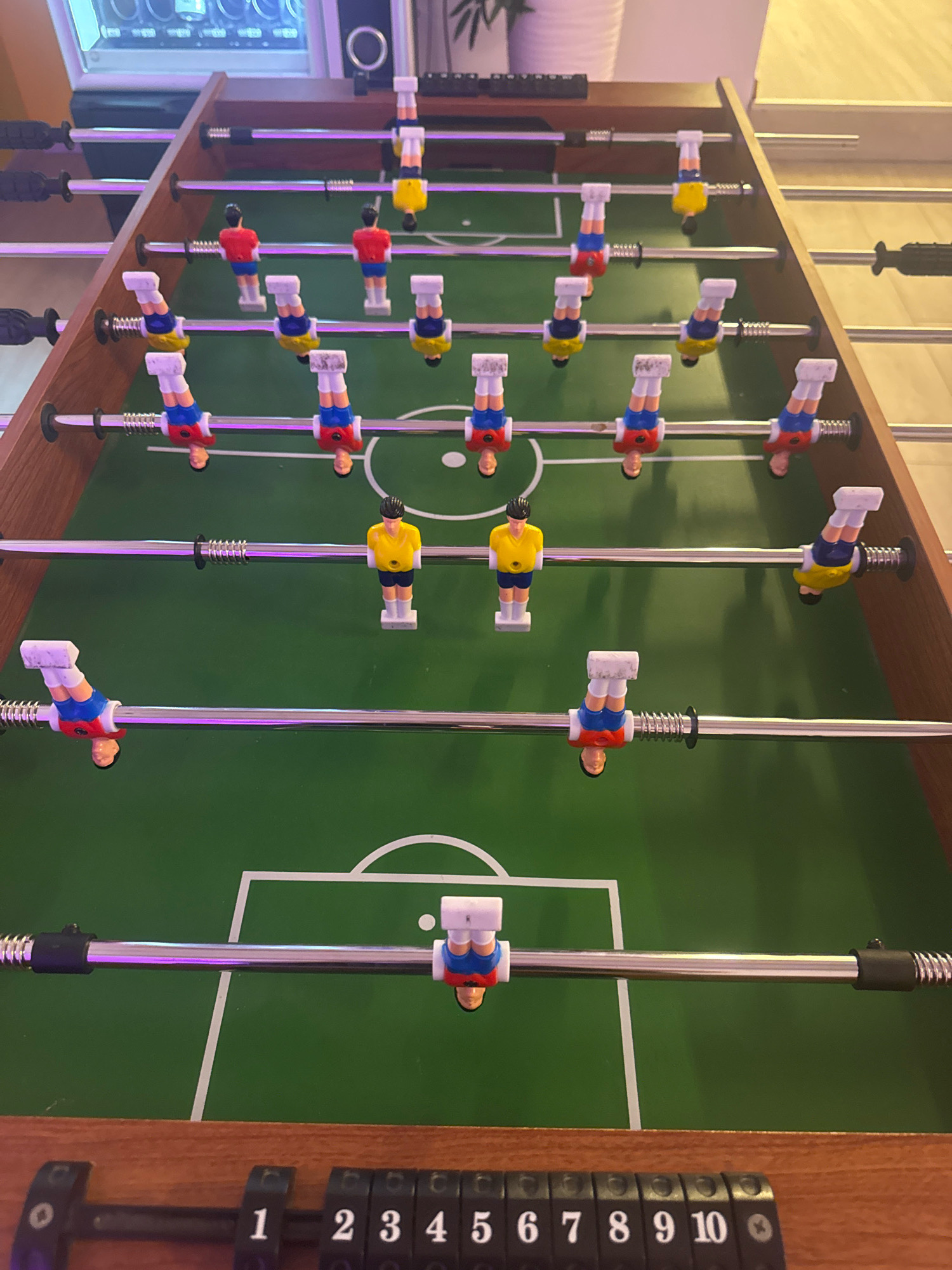 The image shows a foosball table in a dimly lit room. The foosball table has a green playing surface with white lines marking the field. There are several rows of foosball players, some standing upright and some inverted, made of plastic and painted in red, yellow, blue, and white. The players are positioned on metal rods that run across the table. In the foreground, a scoring mechanism with numbered dials (1-10) is visible. The background is out of focus but suggests an indoor setting with some lighting. The exact location in Germasogeia Municipality, Cyprus cannot be determined from the image alone, as it only shows the interior of a room with a foosball table.