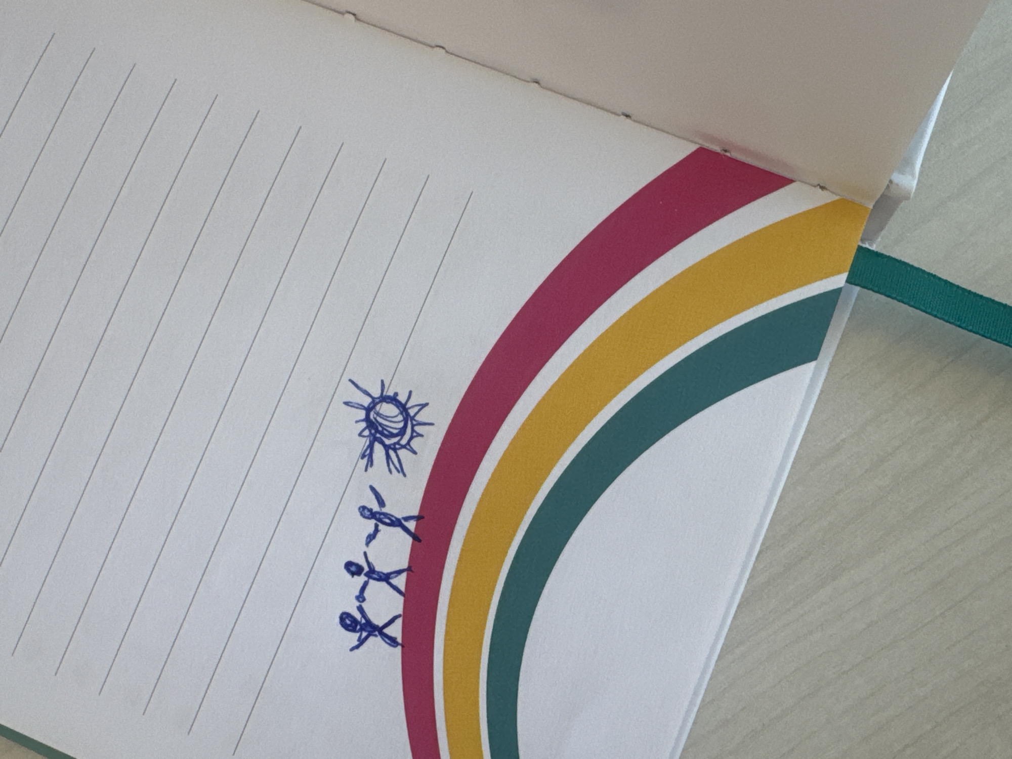 An open notebook lies on a surface, its pages lined and bearing a child-like drawing of the sun and three stick figures. A colorful rainbow design runs along the side of the page. The location is unknown, and there are no indications to show this location is Tel-Aviv, Israel.
