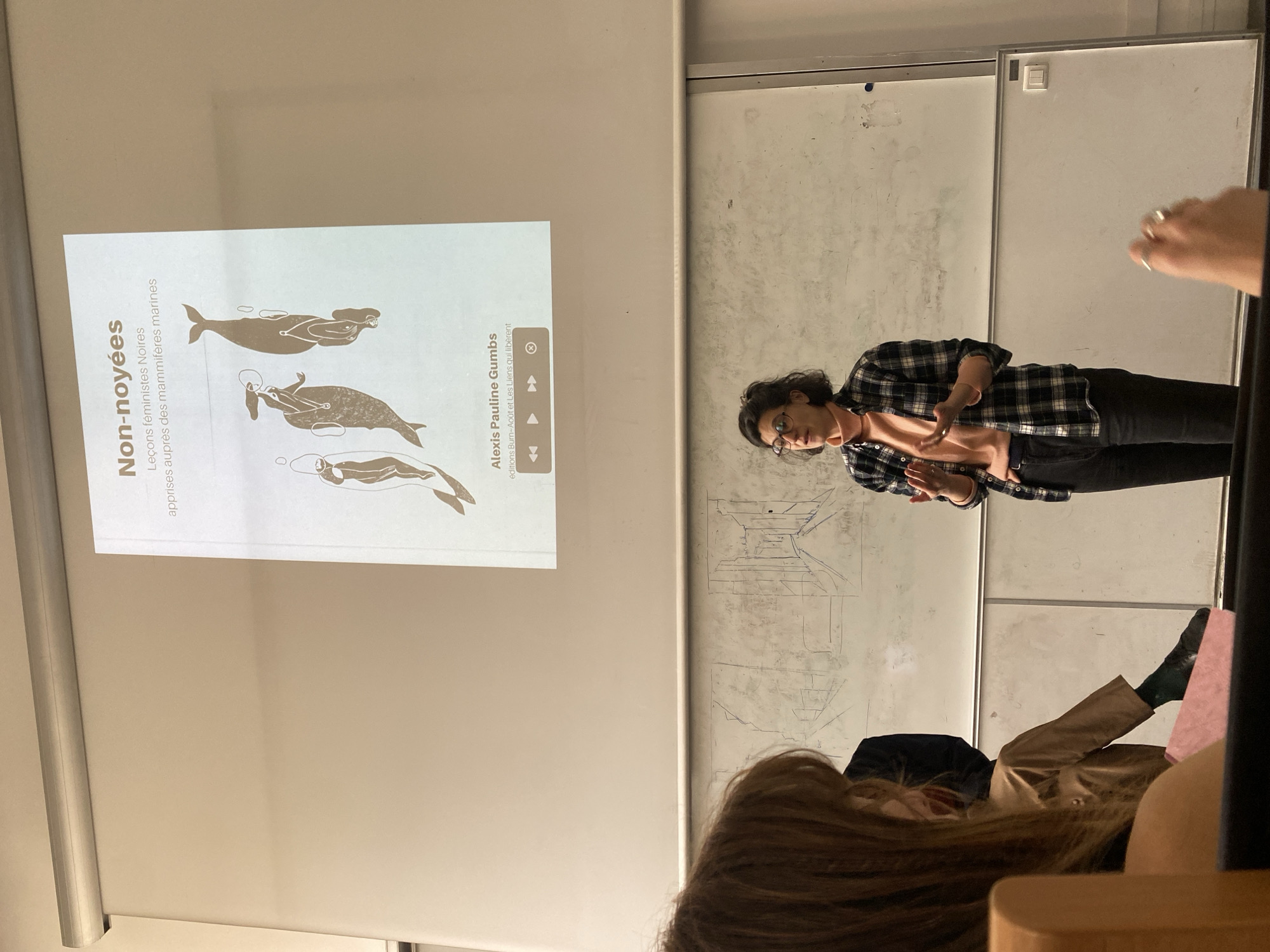 A woman is giving a presentation in a classroom setting, with a projection screen displaying the book cover "Non-noyées," which translates to "Non-drowned" in English, subtitled "Black feminist lessons learned from marine mammals" by Alexis Pauline Gumbs. The woman is dressed in a plaid shirt and glasses, with dark curly hair, standing near a whiteboard with drawings. Seated students are visible in the foreground, including one with long brown hair and another with a tan jacket. The overall environment suggests an educational setting, possibly at a university in Paris, France, given the French language of the book title.