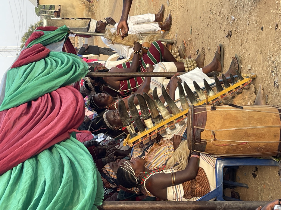 The image captures a vibrant outdoor cultural gathering, presented in a portrait orientation where the subjects appear to be lying on their sides, suggesting the original photo was taken with the camera rotated or the subjects are actually reclining.
In the foreground, taking up the left side of the frame, are large swathes of fabric in bright green and reddish-pink, meticulously draped and folded. These textiles form a colorful, textured backdrop or part of a temporary structure.
Beneath and extending to the right of these fabrics is a diverse group of people, primarily adults with dark complexions, engaged in an event. Many are reclining or lying down on what appears to be a raised platform or ground covering. They are adorned in various traditional or ceremonial garments. Several individuals wear striped fabrics in patterns of red, green, black, and white. Some have elaborate head coverings or intricate hairstyles, and many are decorated with beads and jewelry around their necks and arms.
A prominent musical ensemble is central to the scene. A unique instrument composed of numerous animal horns (at least 10-12) is arranged horizontally on a yellow wooden frame, resembling a type of horn xylophone or panpipe. Next to it, a large, cylindrical wooden drum, crafted from lighter and darker wood sections, rests on a blue plastic chair, ready for use.
Among the people, one individual in the lower right section of the image (when viewed upright) is distinctly visible, reclining with a striking light-colored, possibly braided or wig-like, hairstyle and multiple strands of colorful beads. Further back, another person in a white robe and cap, holding what appears to be a light-colored animal fur or tassel, is visible. The bare feet of several individuals are seen, some wearing sandals, others boots.
The setting appears to be outdoors on dry, light brown ground or dirt. In the background, hints of other structures, possibly buildings, and distant vegetation are visible under what seems to be bright daylight. The gathering, traditional attire, and musical instruments suggest a ceremonial performance, festival, or cultural celebration is taking place. The relaxed, reclining postures might indicate a period of rest during a long event or a customary way of observing the proceedings.