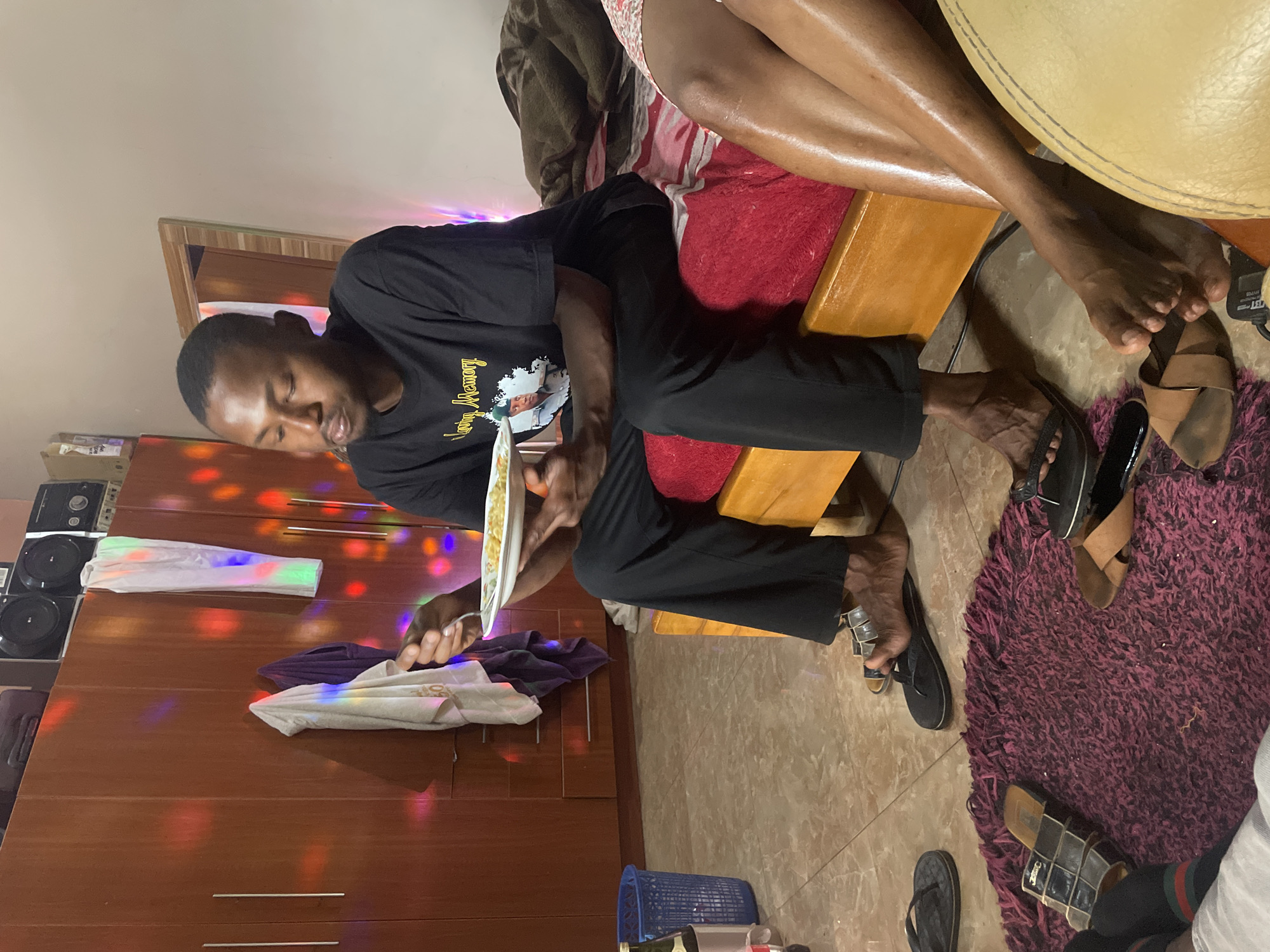 The scene is indoors, likely a residence in Jos, Nigeria. An African man in a black t-shirt is seated, holding a plate of food and a fork. The room features a wooden cabinet with clothing items on top, a red cushion, a round leather stool, and a purple rug. Several pairs of sandals are scattered on the floor. The lighting is colorful, creating a party-like ambiance. Another person's legs are visible, seated on the stool. The man is focused on eating his meal.