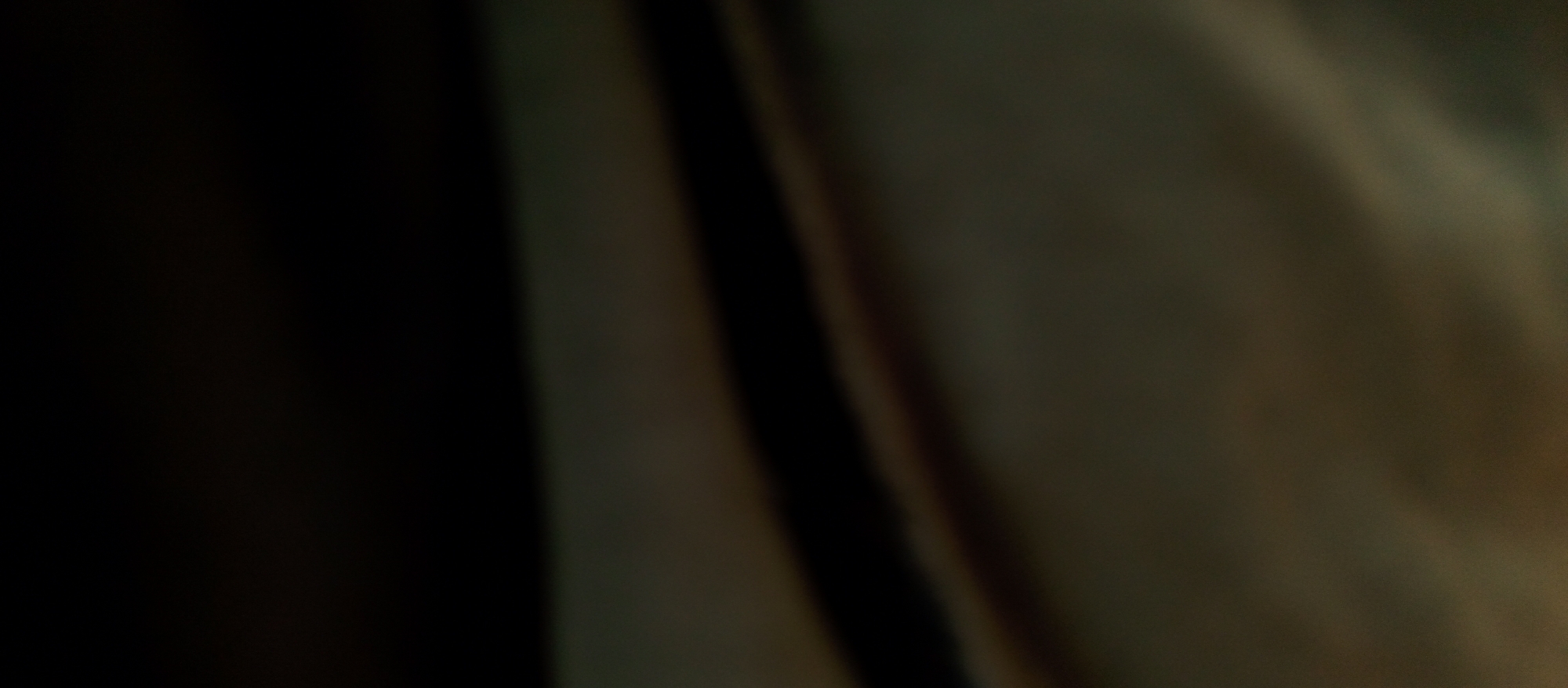 The image presents an abstract, close-up view dominated by dark tones and muted colors, strongly suggesting draped fabric or a textile surface. The left side of the frame is almost entirely black, indicating deep shadow. Moving towards the center-right, the image transitions into shades of dark olive green, brown, and subtle ochre. A prominent, soft-edged vertical dark stripe, likely a fold or crease, runs through the mid-right section. To the right of this stripe, the surface appears slightly lighter, revealing subtle, blurry undulations and variations in hue, consistent with the texture of a soft material. The overall impression is one of deep shadow and soft light interplay on an indistinct, textured surface in very low light conditions. There are no discernible people, specific objects beyond the assumed fabric, or identifiable activities taking place. No specific time of day, weather, or explicit location cues are visible within the image content, and no text is present.