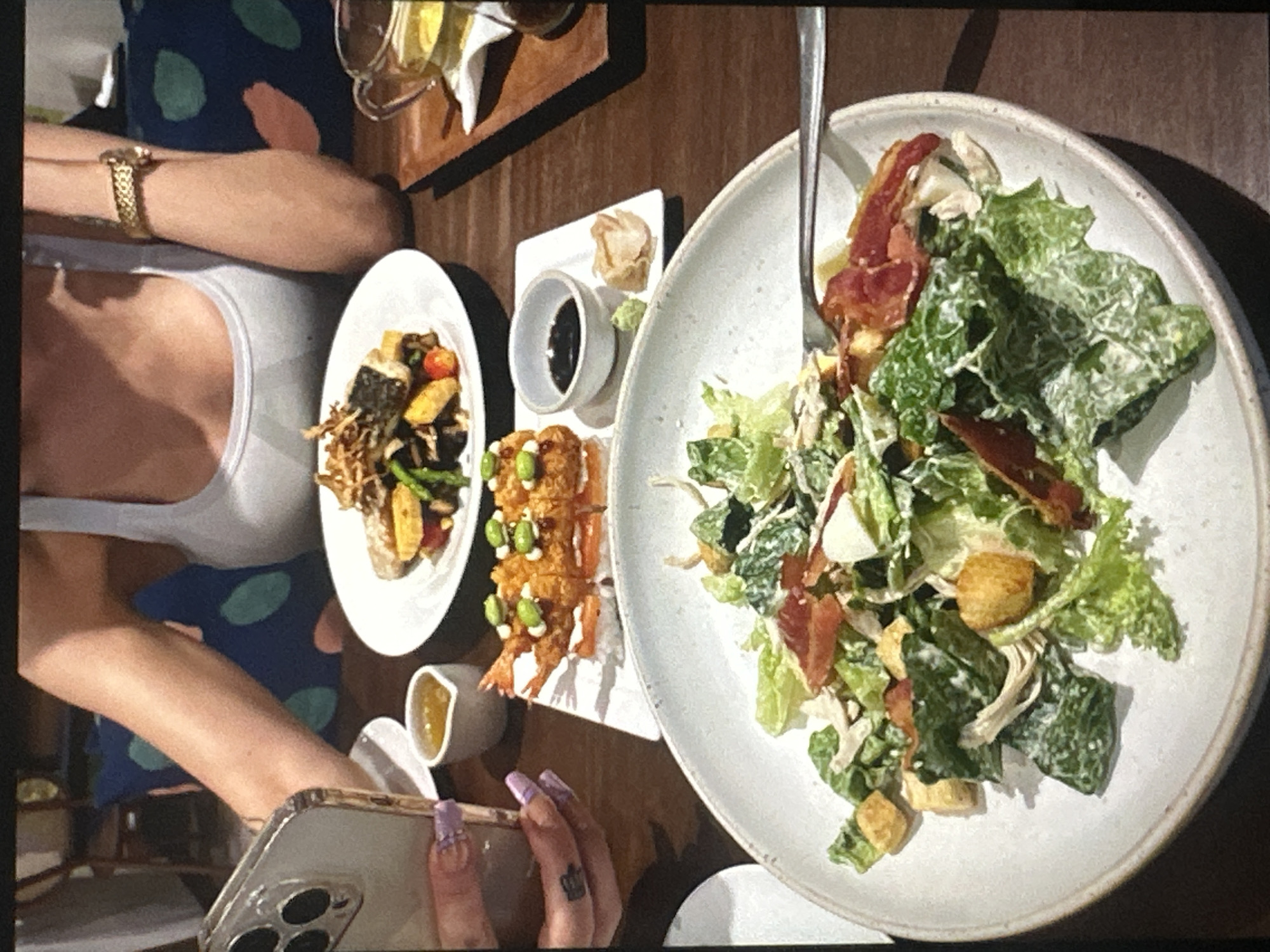 The image shows a table setting with several plates of food, including a salad, fried shrimp with edamame, and a plate of fish and vegetables. A person is sitting at the table, visible from the chest up, wearing a white tank top. Their hands are visible, holding a phone. A glass with a drink is also on the table. The location is unknown.