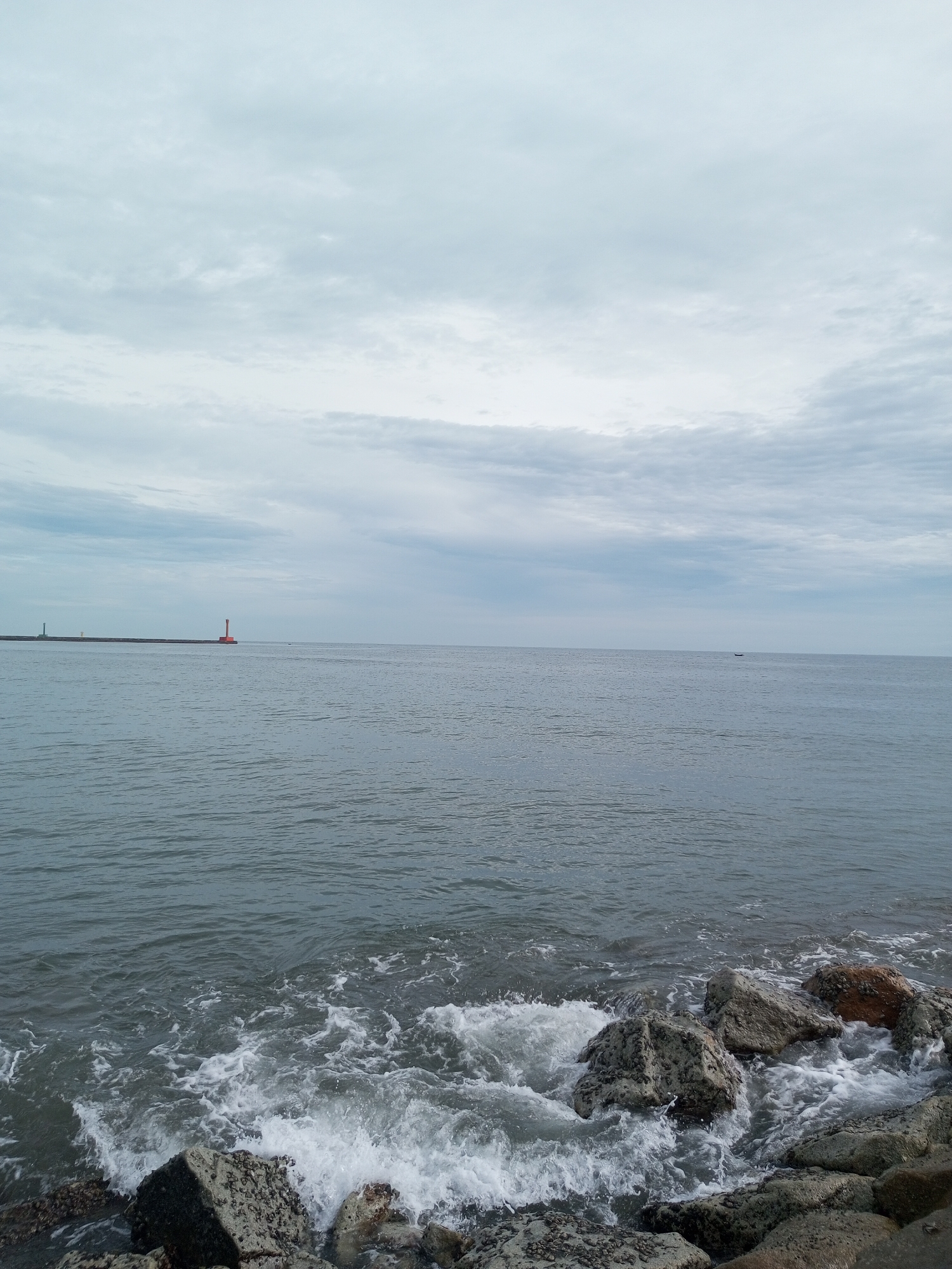 An overcast day at the ocean. The foreground features a rocky shore with waves crashing against the stones, creating white foam. The sea stretches out to the horizon, appearing calm with gentle ripples. In the distance, a long pier or breakwater extends into the water, with two lighthouses visible on it. One lighthouse is red and the other is a lighter color, possibly white or green. A small boat is also visible in the far distance on the water. The sky is filled with clouds, creating a muted and soft lighting condition. The overall scene depicts a coastal environment under a cloudy sky.