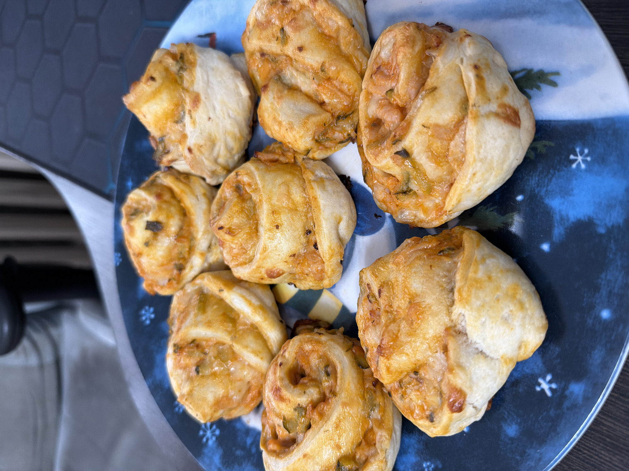 A close-up shot shows a dark blue plate with a festive, snowflake-patterned rim holding eight savory pastry rolls. The rolls, baked to a golden-brown perfection, appear to be filled with a mixture of cheese and vegetables, possibly pizza-flavored. They are arranged in a circular pattern on the plate, with some overlapping. The plate rests on a dark, wooden surface. The background is blurred, but hints at a domestic setting with neutral-colored furniture and a dark object that may be a chair. The image does not provide any information about the city of Chaikovsky, Russia.