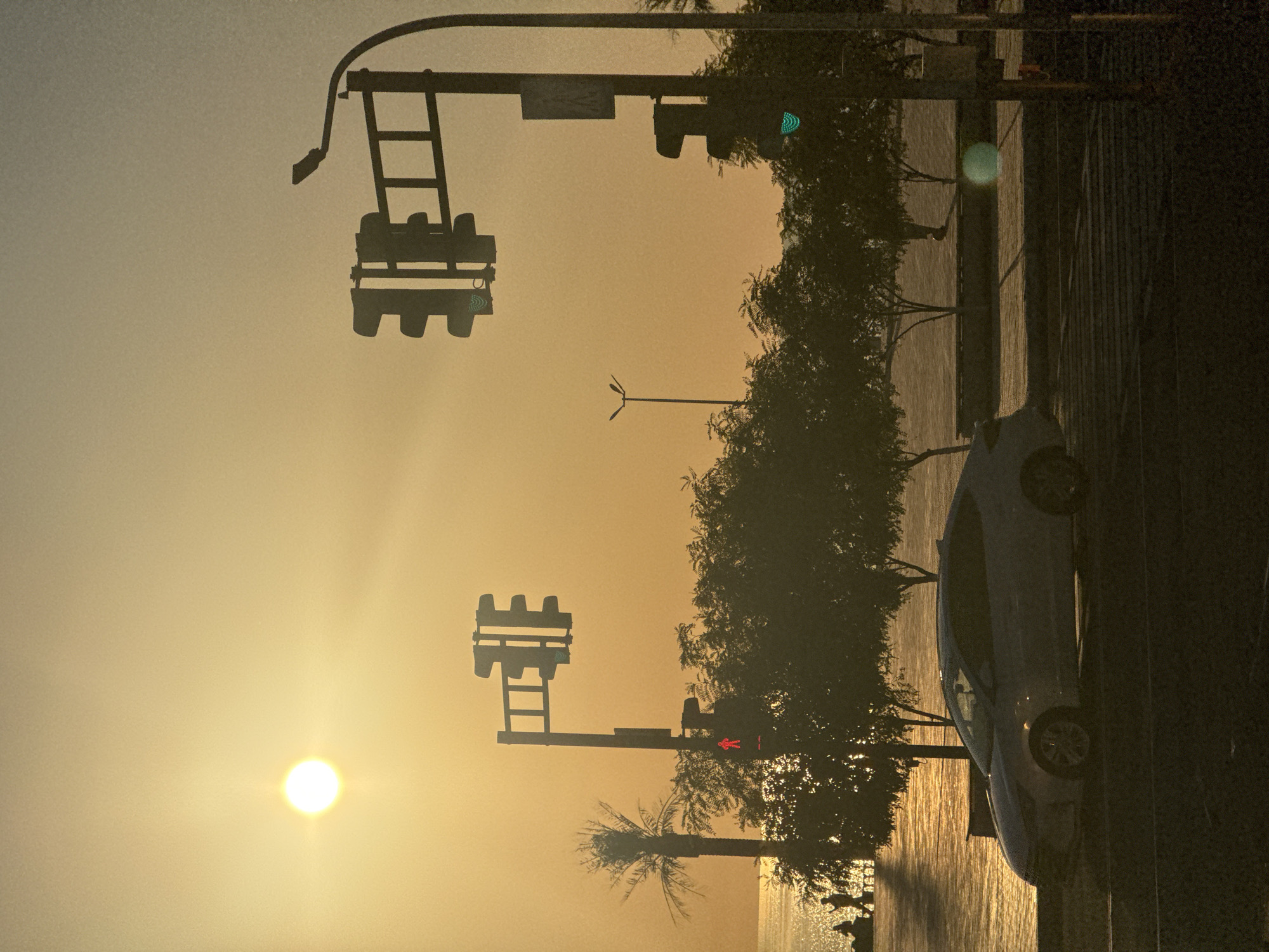 The image depicts a street scene in Aktau, Kazakhstan, at sunset. The sun is low on the horizon, casting a warm, orange glow over the sky and reflecting on the calm body of water in the background, likely the Caspian Sea. In the foreground, a white car is parked or stopped near a street intersection. Traffic lights are visible, with one showing a red pedestrian signal and another showing a green traffic light. Silhouetted trees line the waterfront, and a palm tree stands to the left. Several indistinct figures are seen walking along the waterfront promenade. The overall atmosphere is tranquil, with the warm light and reflections creating a serene ambiance.
