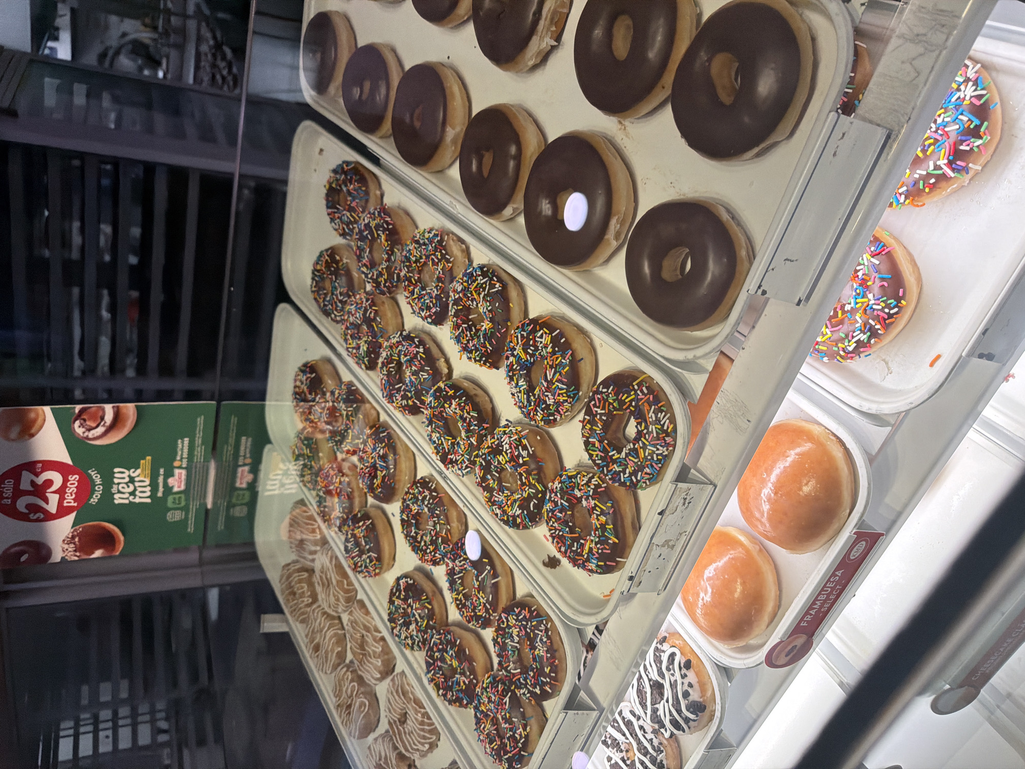 This image captures a static, brightly lit display case filled with a variety of donuts and pastries inside a commercial establishment, likely a donut shop or bakery, located in San Bernardino Tlaxcalancingo, Mexico.
The display case features multiple white rectangular trays arranged in rows. The trays contain numerous donuts with diverse toppings:
* Several trays are filled with chocolate-glazed donuts liberally coated with colorful sprinkles.
* Adjacent trays hold plain dark chocolate-glazed donuts, some exhibiting a small, reflective white circular mark on their surface.
* In the upper left section, a tray displays light-colored donuts adorned with intricate white icing drizzles and patterns.
* Below, two large, round, un-holed pastries with a glossy, orange-pink glaze are present, accompanied by a label reading "FRAMBUESA - SELECT KMD," indicating a raspberry selection.
* At the bottom right, two sliced half-donuts, glazed and covered with colorful sprinkles, are visible, exposing their cut interiors.
* A partially visible tray towards the bottom left contains both elongated, rectangular donuts and round donuts, all decorated with alternating dark and white icing drizzles.
A green promotional sign is prominently displayed in the upper right quadrant, featuring red circular text that states "a solo $23 pesos SOLO HOY! the new favs" (only $23 pesos TODAY ONLY! the new favs), indicating a special offer. Subtle branding, including what appears to be a Krispy Kreme logo, is visible on this sign. The clear glass of the display case reflects ambient light and portions of the establishment's interior. The background visible through the glass consists of dark, horizontal elements resembling shelving or storage racks. No individuals are present in the frame, and no actions or interactions are depicted.