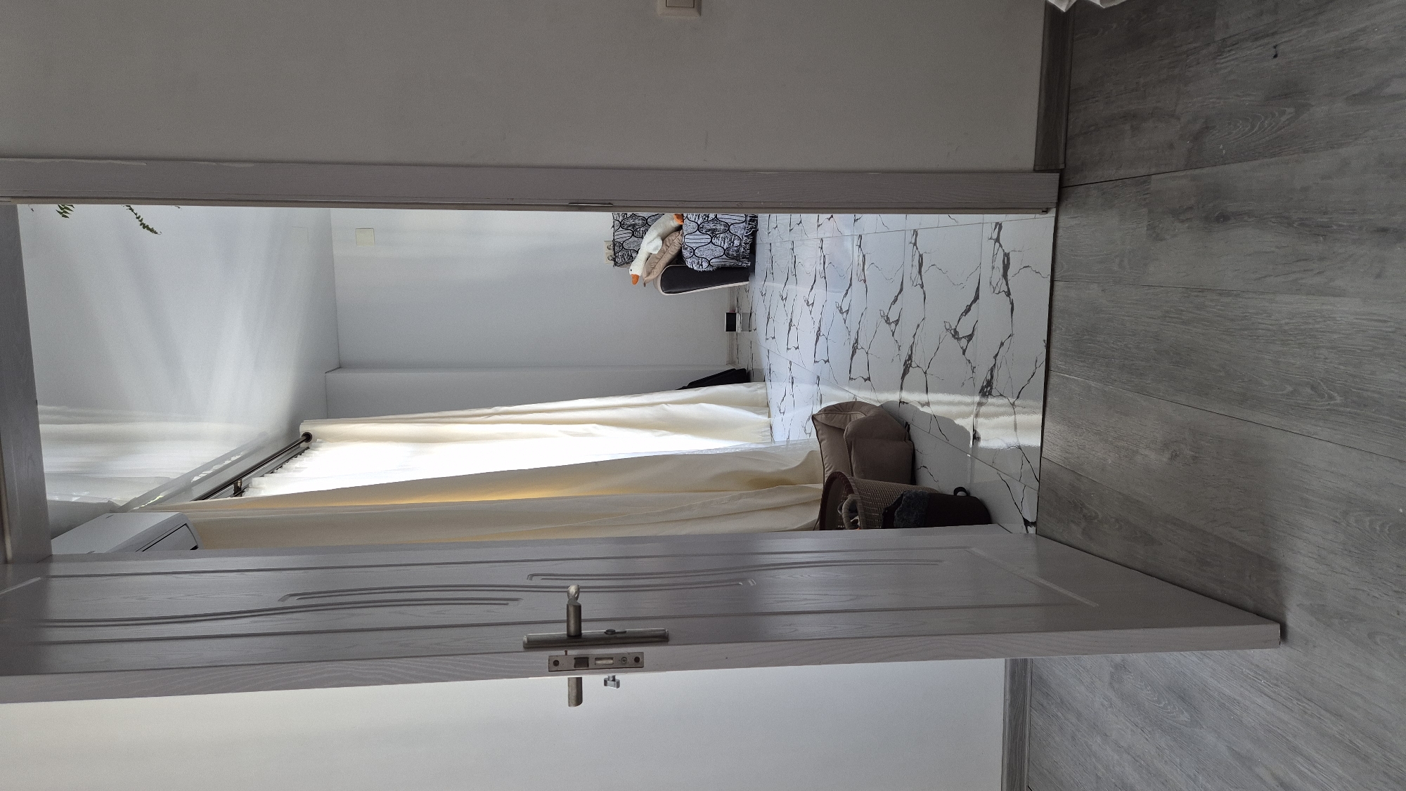 The photograph depicts a room in Batumi, Georgia, as viewed from an open doorway. Inside, the walls are painted white. A white curtain hangs across the far wall, partially obscuring what appears to be an air conditioning unit mounted below the curtain rod. The floor is covered in white marble tile with dark gray veining. There is a small chair with a patterned seat. A stuffed toy is on the chair. Several soft bags are also on the floor. Outside the room, the flooring is gray wood-look laminate. The door is painted white with decorative grooved panels. A simple silver door handle and latch mechanism are visible.