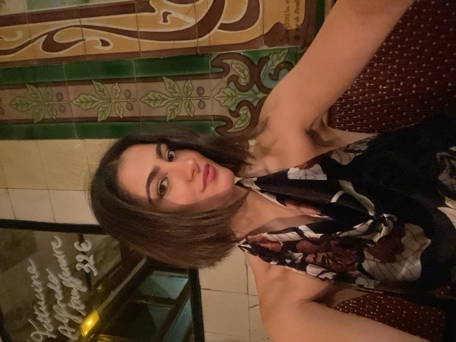 A woman with shoulder-length brunette hair and fair skin poses indoors. She is wearing a floral print top, seated against a wicker patterned backdrop. A menu board with handwritten French script and a price of 32€ is visible to her left. Decorated tile work with green, brown, and beige tones lines the wall behind her, creating a patterned background. The scene has warm, dim lighting.