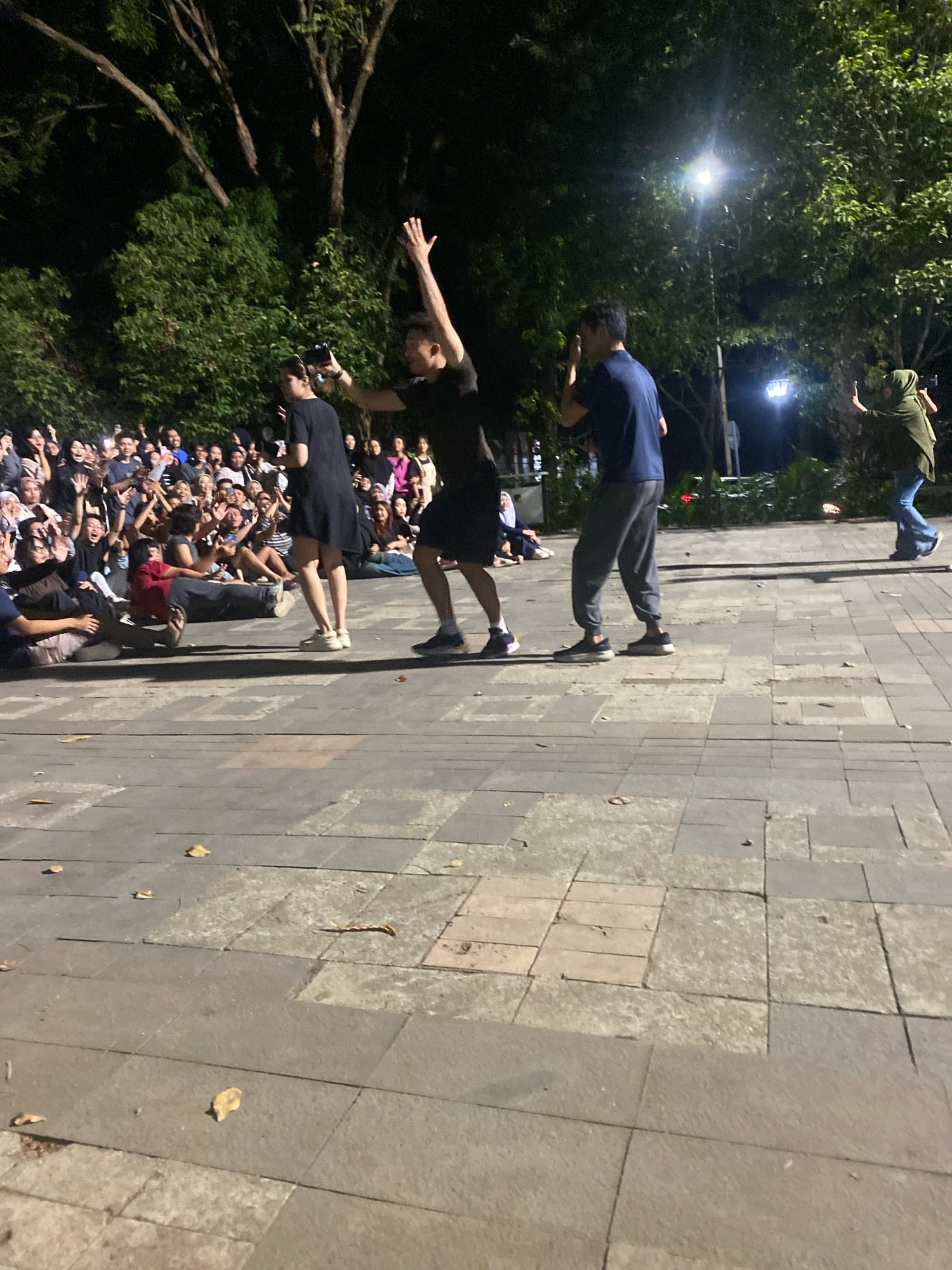 At night, a group of people gathers on a stone-paved walkway in Semarang, Indonesia, seemingly watching a performance or event. In the foreground, the walkway is composed of large square stone tiles, some of which are uneven or discolored, with a few fallen leaves scattered across them. Behind the walkway, a dense crowd of onlookers, most of whom are young adults, are visible, many with their arms raised and phones out, suggesting they are recording or taking pictures.
The performers are in the center of the scene. One woman in a black dress is holding a phone and appears to be speaking or singing. To her right, a man in a black t-shirt and shorts is energetically gesturing with his arms raised. Further to the right, a man in a blue t-shirt and grey sweatpants is also raising his hand, facing the crowd. Beyond the performers, trees and greenery form a dark backdrop, with streetlights providing some illumination. The overall atmosphere is one of lively engagement and community gathering.