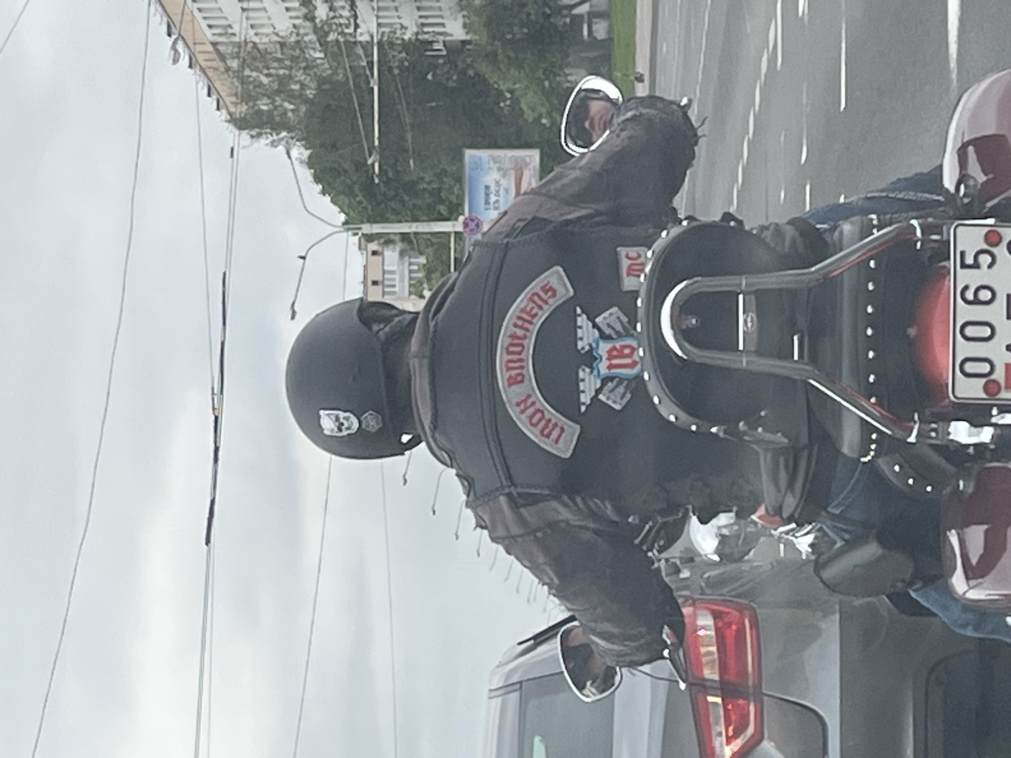 The image, taken from a slightly elevated perspective and rotated 90 degrees clockwise, captures a person on a motorcycle from behind, seemingly stationary in urban traffic. The motorcyclist wears a dark leather jacket with several patches. A prominent patch on the back displays the words "IRON BROTHERS" in an arc, above a stylized "IB" logo, and a smaller patch with "MC" below. The rider's black helmet features a white symbol on the back, possibly a skull, with partially visible letters "DO" or "D0".
The motorcycle has a large, studded seat and a chrome backrest. Its license plate is partially visible, showing "0065 AZ", which is consistent with Belarusian registration, given the location context of Minsk, Belarus. Behind the motorcycle, the front of a grey car is visible. In the background, there are blurred buildings, green trees, and overhead power lines against an overcast sky, indicating daytime. A billboard with unreadable Cyrillic text can be seen in the distance. A faint reflection or another person is visible in the motorcycle's right rearview mirror.