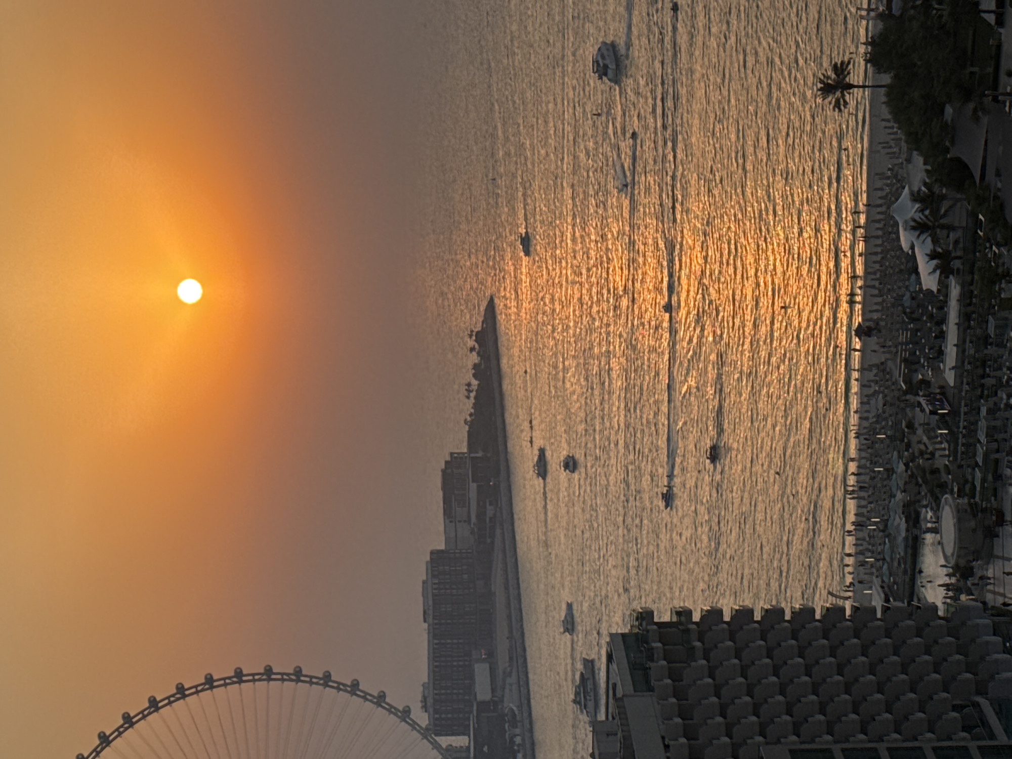 This is a high-angle, full-frame, vertical, daylight, medium shot photograph taken in Dubai, United Arab Emirates, capturing a sunset over the Arabian Gulf. The sun, a bright white orb, is positioned slightly right of center in the upper half of the frame, casting a warm, orange glow across the sky and reflecting intensely on the water's surface.
To the left of the frame, a large Ferris wheel, identified as Ain Dubai, is partially visible, its structure silhouetted against the sunset. In the middle ground, a long, low-rise urban development stretches along the coastline, featuring multi-story buildings with dark facades and numerous windows. A body of water separates this development from the foreground.
The water is populated by several boats, including a white motor yacht in the middle left, and various smaller vessels and personal watercraft. Their wakes are visible, creating lines of disturbance on the shimmering water.
The foreground is dominated by the facade of a large, modern building with a distinctive geometric pattern of balconies, occupying the lower left quadrant. Below this building, a busy beach is visible, lined with rows of white umbrellas and sun loungers. Numerous people are scattered across the beach, some walking, others appearing to be sunbathing or swimming. The water near the beach is calmer, with a few individuals visible swimming. Palm trees and dense foliage dot the edge of the beach and the areas behind it, suggesting a resort or recreational area.
The overall atmosphere is one of a serene, yet active, coastal evening in a major urban center. The hazy effect in the sky suggests either atmospheric dust or the soft light of sunset.