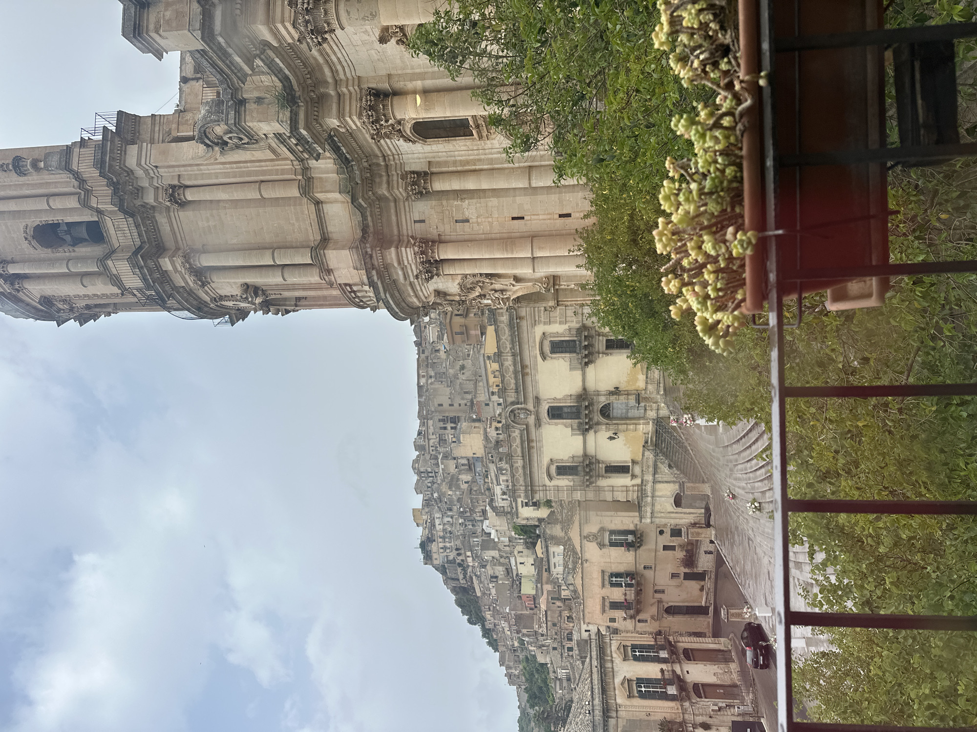 The image shows a view in Modica, Italy, dominated by the architecture and urban landscape. The upper left of the image is filled by a tall, ornate building with intricate details and multiple levels. The architecture is characterized by columns and decorative elements. In the middle, the cityscape cascades down the hillside, composed of numerous buildings tightly clustered together. The buildings vary in color, size, and style. On the right side of the frame, a section of a balcony is visible, including a railing and a flower pot filled with foliage. The sky is overcast.