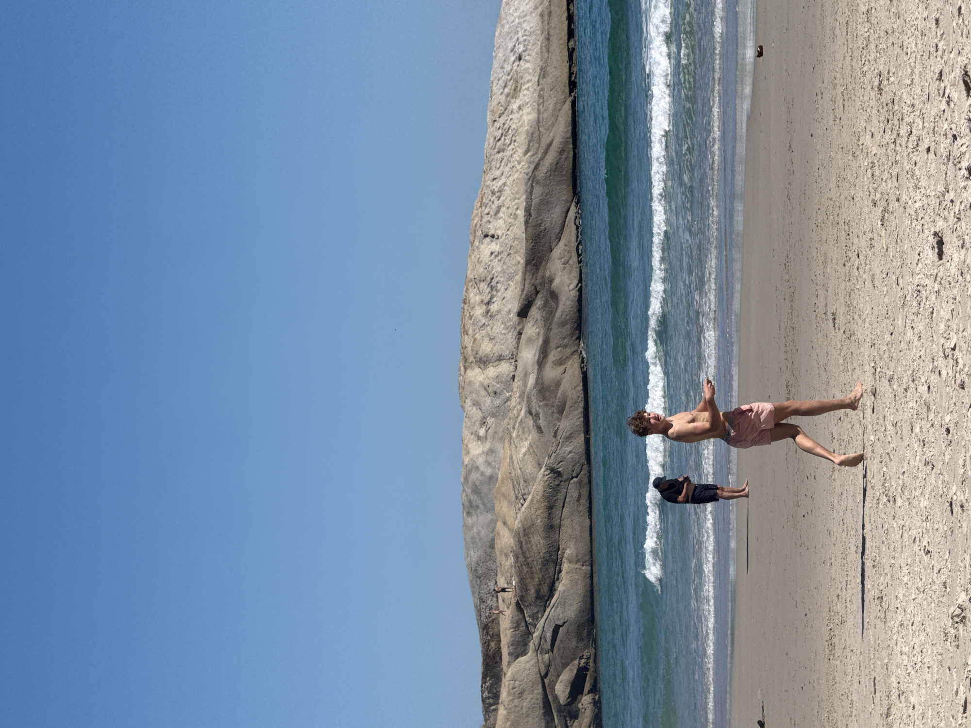 The image shows a vertical view of a beach scene in Hout Bay, South Africa. The sky is clear and blue. A large, light-colored rock formation runs vertically along the mid-left of the image. The ocean with small white waves is situated next to the rock formation. Two people are standing on the beach: one is wearing pink shorts, has bare chest and face turned towards the ocean. The other person is wearing dark shorts and a dark shirt and is also facing the water. The sand is pale in color.