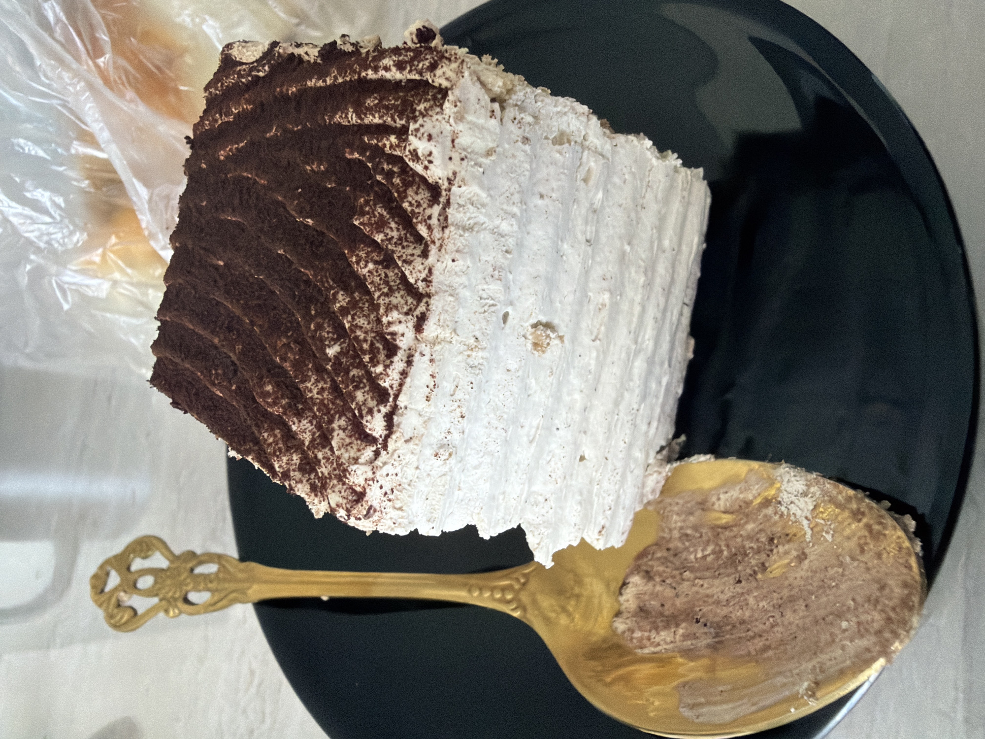 A slice of layered cake, dusted with cocoa powder on top and white frosting in between the layers, sits on a black plate. A decorative gold spoon with some cake residue on it rests to the left of the cake. The background is blurred, showing a white surface and a clear plastic bag with some baked goods inside. This image was taken on September 11, 2023, at 11:55 AM in Strășeni, Moldova.