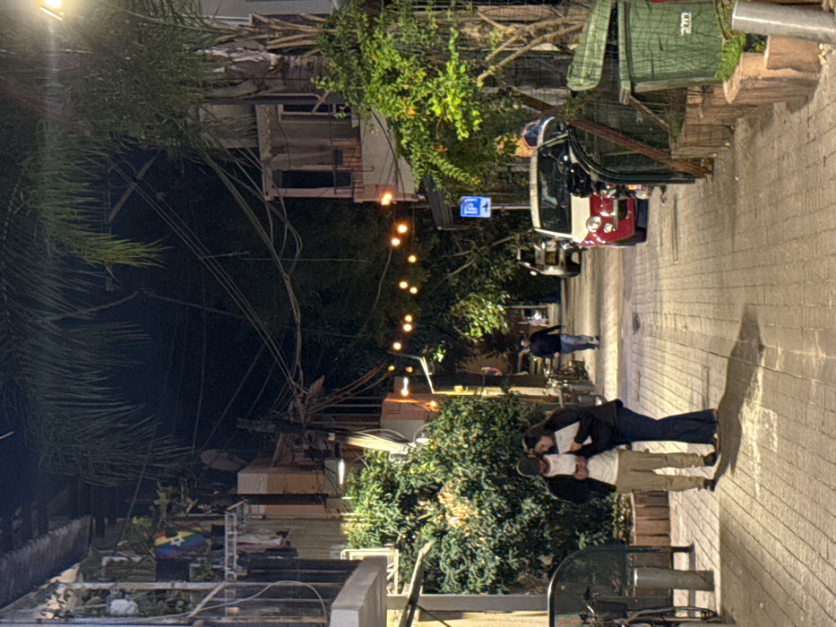 A nighttime street scene features a couple embracing on a brick-paved sidewalk. Above, strings of lights illuminate the path. A vehicle is parked nearby, with a green bin visible behind it. A person walks further down the street. Buildings line the left side, partially obscured by trees and utility lines.