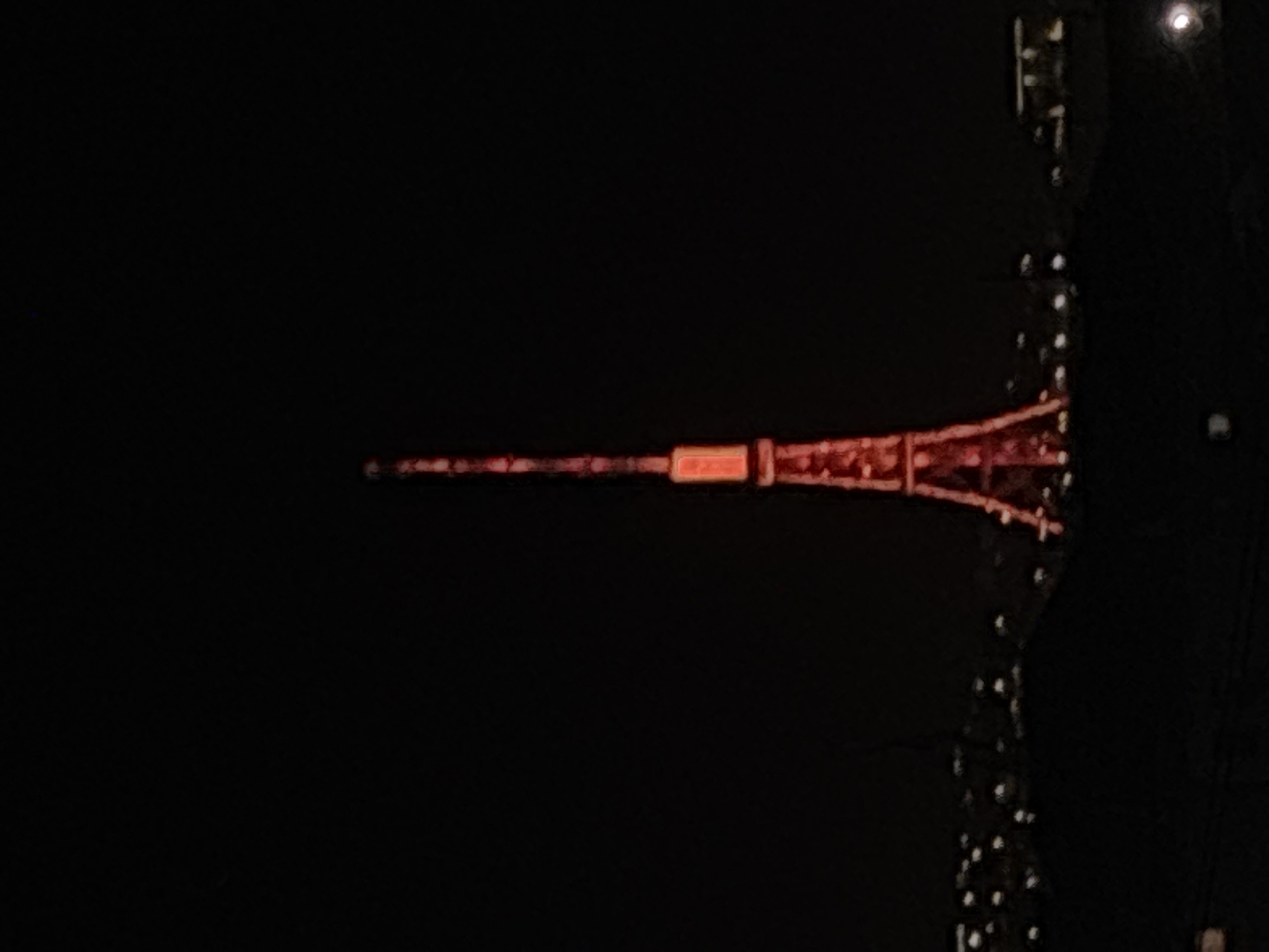 The image shows the Yerevan TV Tower at night. The tower is illuminated and visible against a dark sky. The surrounding environment is obscured by darkness, but there are some lights and structures near the tower.