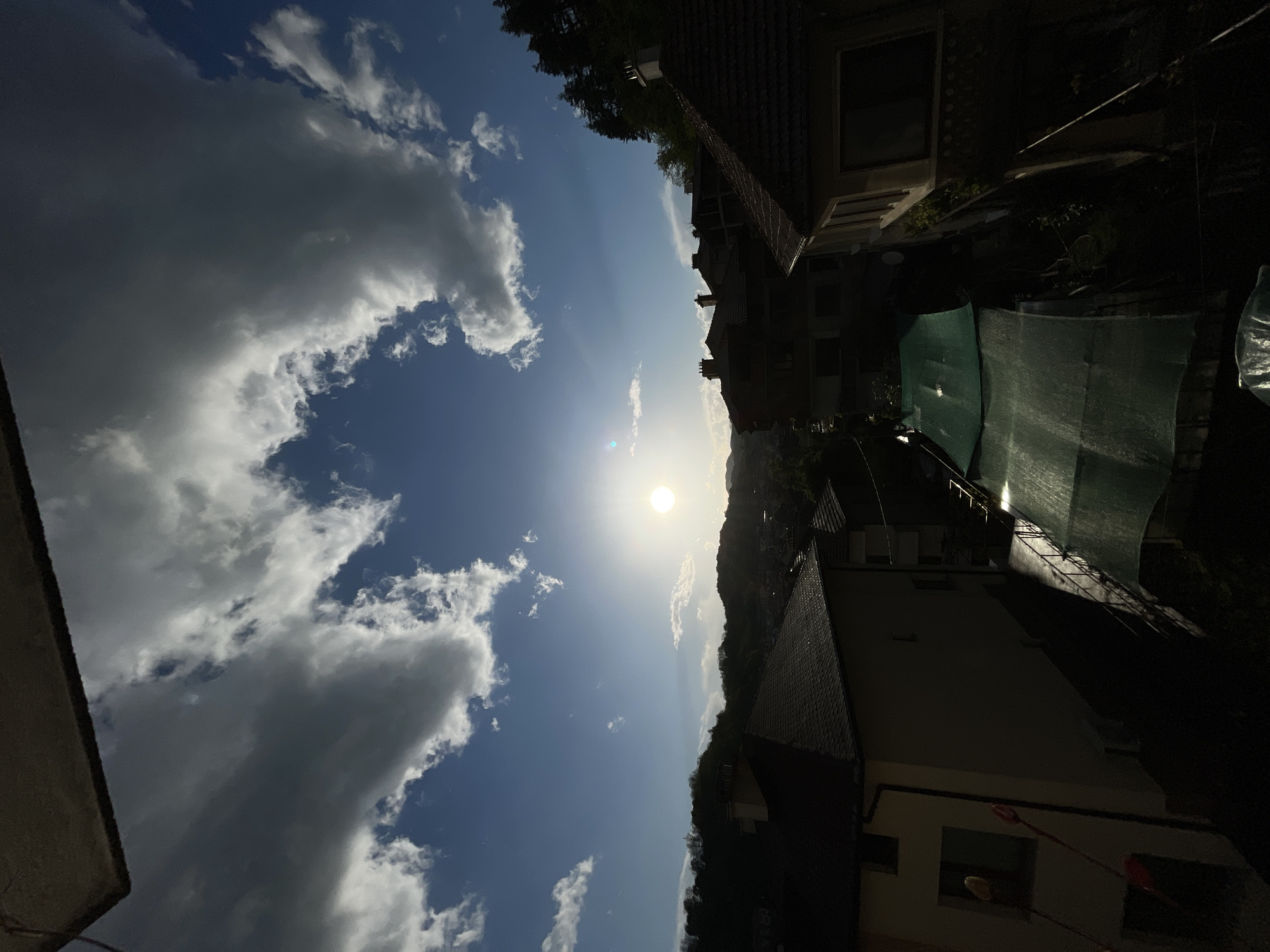The image shows a daytime scene, likely in Zlatograd, Bulgaria, though specific confirmation from the image alone is absent. The sky is bright blue, partially obscured by large, fluffy white clouds. The sun is visible and intense, positioned in the center of the image.
On the right side of the image are densely packed residential buildings, appearing dark due to the strong backlight. One building is partially covered by a green mesh or tarpaulin. The terrain appears hilly or mountainous in the background.
