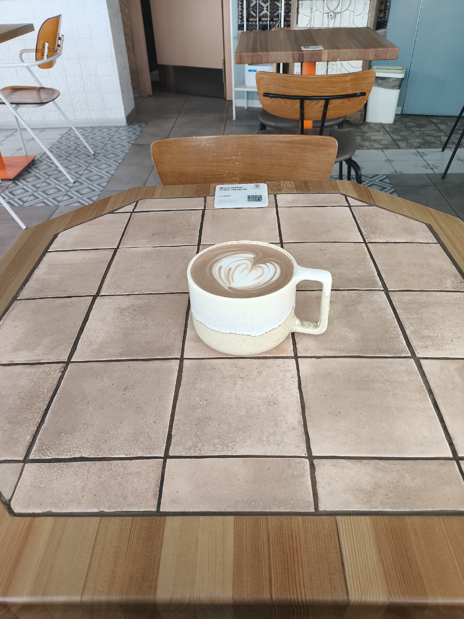 The image shows a close-up of a mug of hot chocolate with latte art on a tiled tabletop in what appears to be a café setting. The tabletop is made of square tiles with dark grout, surrounded by a wooden edge. A wooden chair is visible in the background. Other chairs and tables can be seen in the blurred background, including one with an orange base. The overall environment suggests a casual or modern café in a city of Unknown, Unknown.
