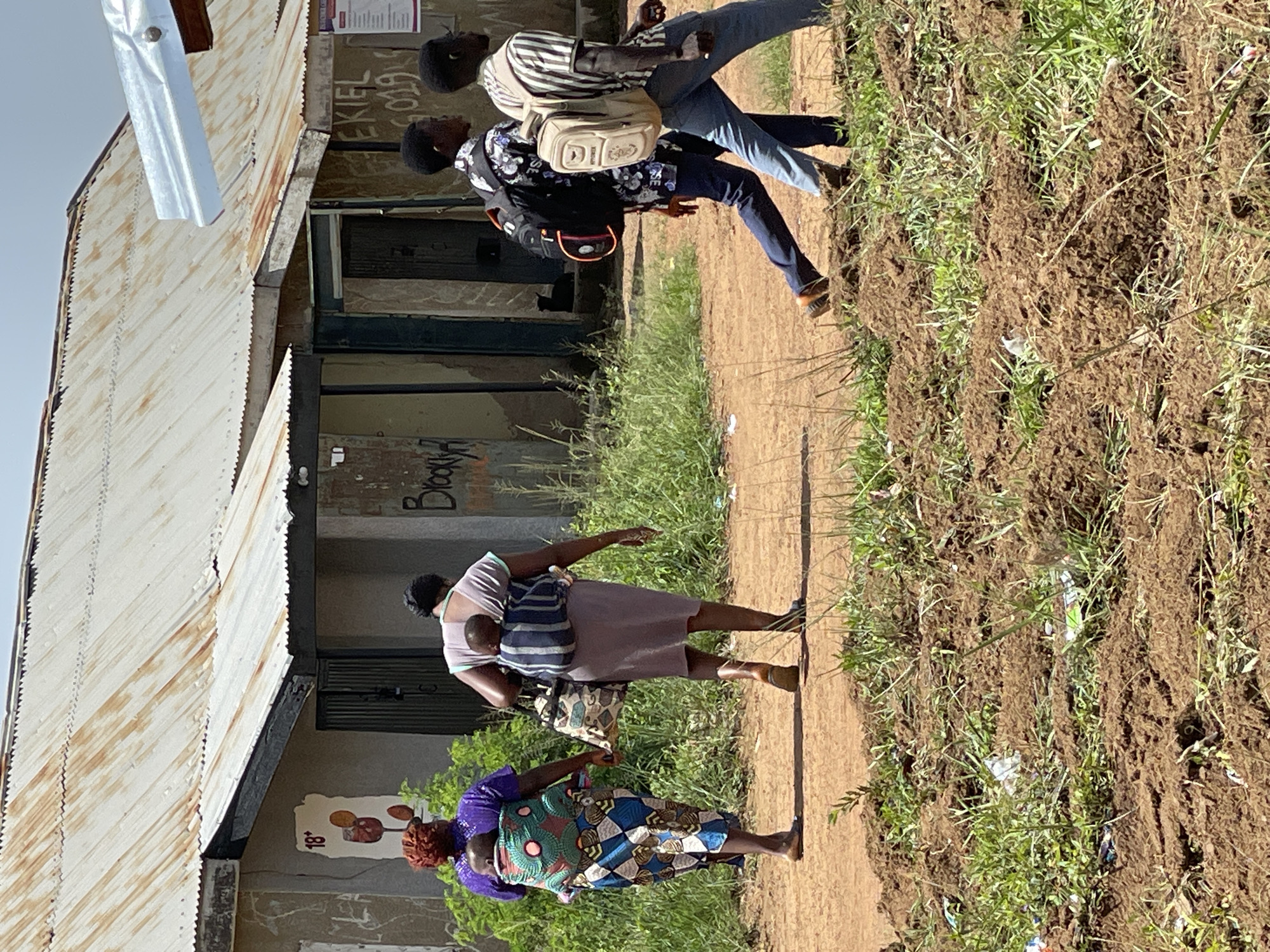 In the city of Durumi, Nigeria, two women are walking, each carrying a child on their back. The woman on the left wears a purple top and a patterned skirt. The woman on the right wears a light-colored dress and carries a bag. Behind them, two young men are walking, one with a black backpack and the other with a beige backpack. They are all walking on a dirt path in front of a row of buildings with corrugated metal roofs. The buildings appear to be somewhat dilapidated, with graffiti on some of the walls. The area is overgrown with grass and dry vegetation. The sky is clear and blue.