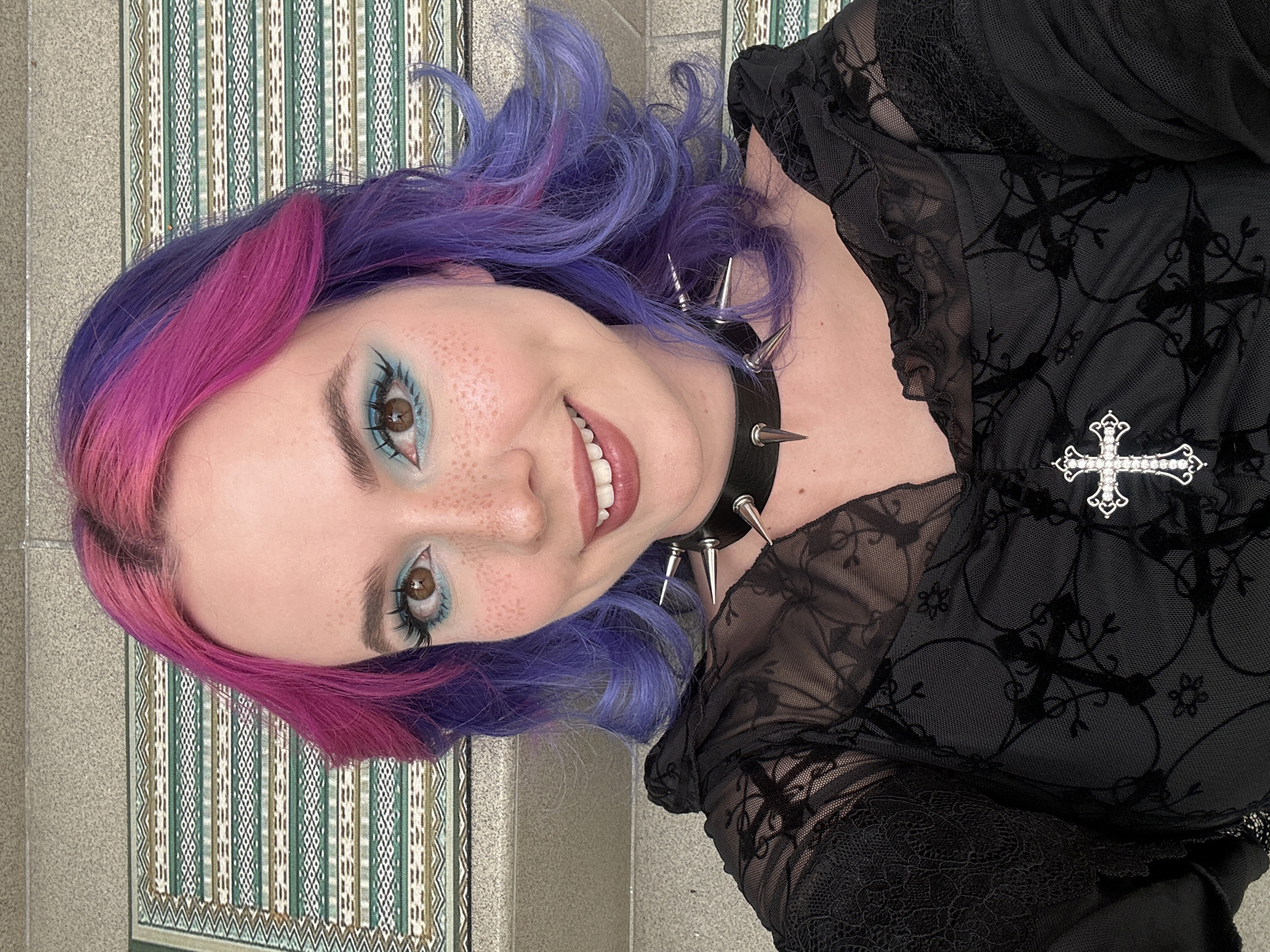 The central subject is a young person smiling directly at the viewer. They have distinct two-toned hair, with bright pink on one side and vibrant purple on the other, styled in loose waves. Their light complexion is dotted with freckles. Elaborate eye makeup features bright teal eyeshadow on the lids and lower lash line, complemented by dark eyeliner and heavy mascara, highlighting their brown eyes. A broad smile reveals white teeth.
They wear a black choker adorned with silver spikes. Their attire includes a black top made of sheer and lace fabric, patterned with flocked crosses and leafy designs. A silver-toned, ornate cross brooch embellished with clear rhinestones is pinned to the top.
The background presents an indoor setting, likely a wall or floor, partially covered by a patterned textile. This textile exhibits vertical stripes in shades of green, white, and brown, incorporating geometric motifs. The overall impression is a cheerful portrait with an alternative fashion aesthetic. No text is visible in the image.