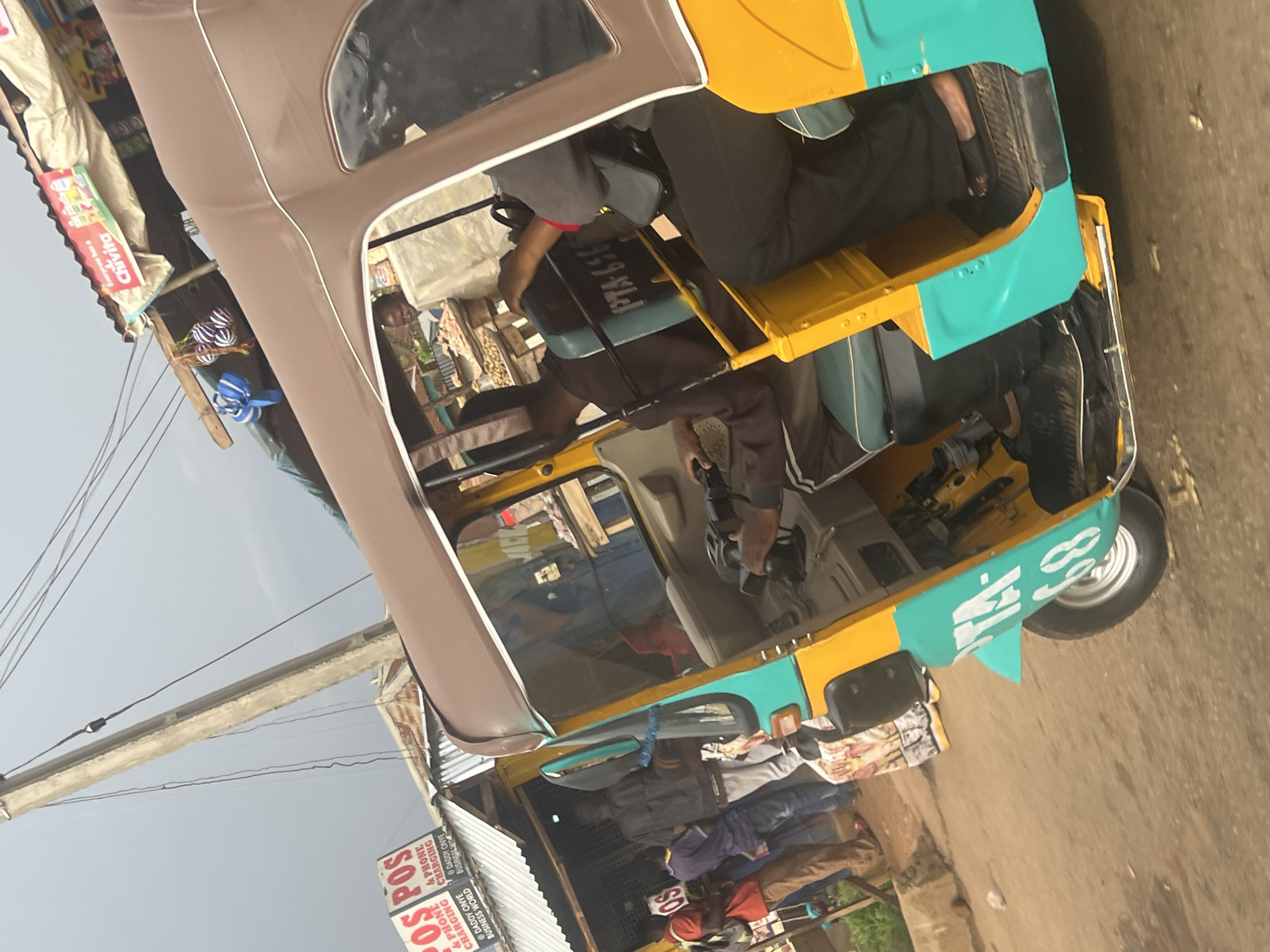 The image depicts a scene in Bukuru, Nigeria, focusing on a yellow and teal auto-rickshaw parked on a dirt road. The rickshaw has a brown roof and several passengers visible inside. In the background, there are stalls with various goods, including what appear to be food items and beverages. One stall prominently displays a sign for "Chivita" juice. Several people are standing near the stalls, some appearing to be customers or vendors. A utility pole with electrical wires stretches across the sky, indicating an urban or semi-urban environment. The overall atmosphere suggests a bustling local market or roadside commercial area. The auto-rickshaw has "PTA 658" written on its side.