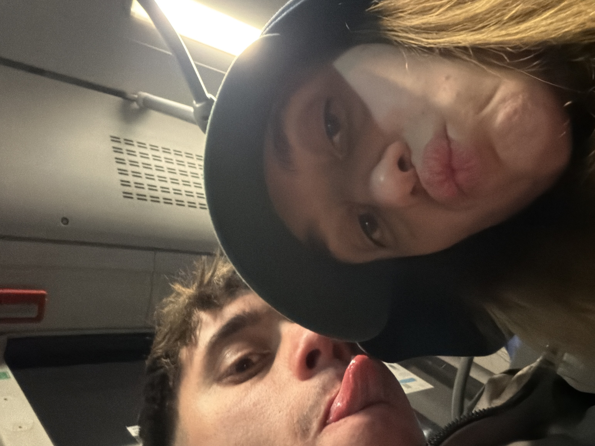 A close-up selfie captured on a mode of public transportation, potentially a bus or train, in an unknown location. Two individuals are visible. One, possibly female, has long blonde hair and is wearing a dark-colored hooded jacket. They are puckering their lips for the camera. The other individual, possibly male, has short brown hair and is sticking their tongue out. The background includes what appears to be the interior of a public transit vehicle, including a vent and a handrail. The exact location within Sofia, Bulgaria, cannot be determined from the image.