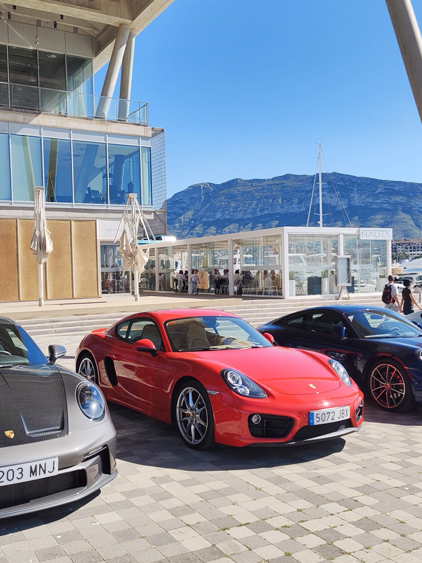 The image shows a bright sunny day in Dénia, Spain, featuring three Porsche cars parked near a waterfront area. The cars, from left to right, are gray, red, and dark blue, with their respective license plates partially visible. The red car's license plate is "5072 JBX". The cars are parked on a paved area.
In the background, a modern building with large glass windows and a tan facade stands. A sign reading "NAUTA" is visible on a nearby glass structure, suggesting a restaurant or similar establishment. Several people are seated at tables inside, while others are walking nearby.
In the distance, a mountain range is visible under a clear blue sky. A sailboat mast is also visible. The overall scene suggests a vibrant, upscale location in the city of Dénia.