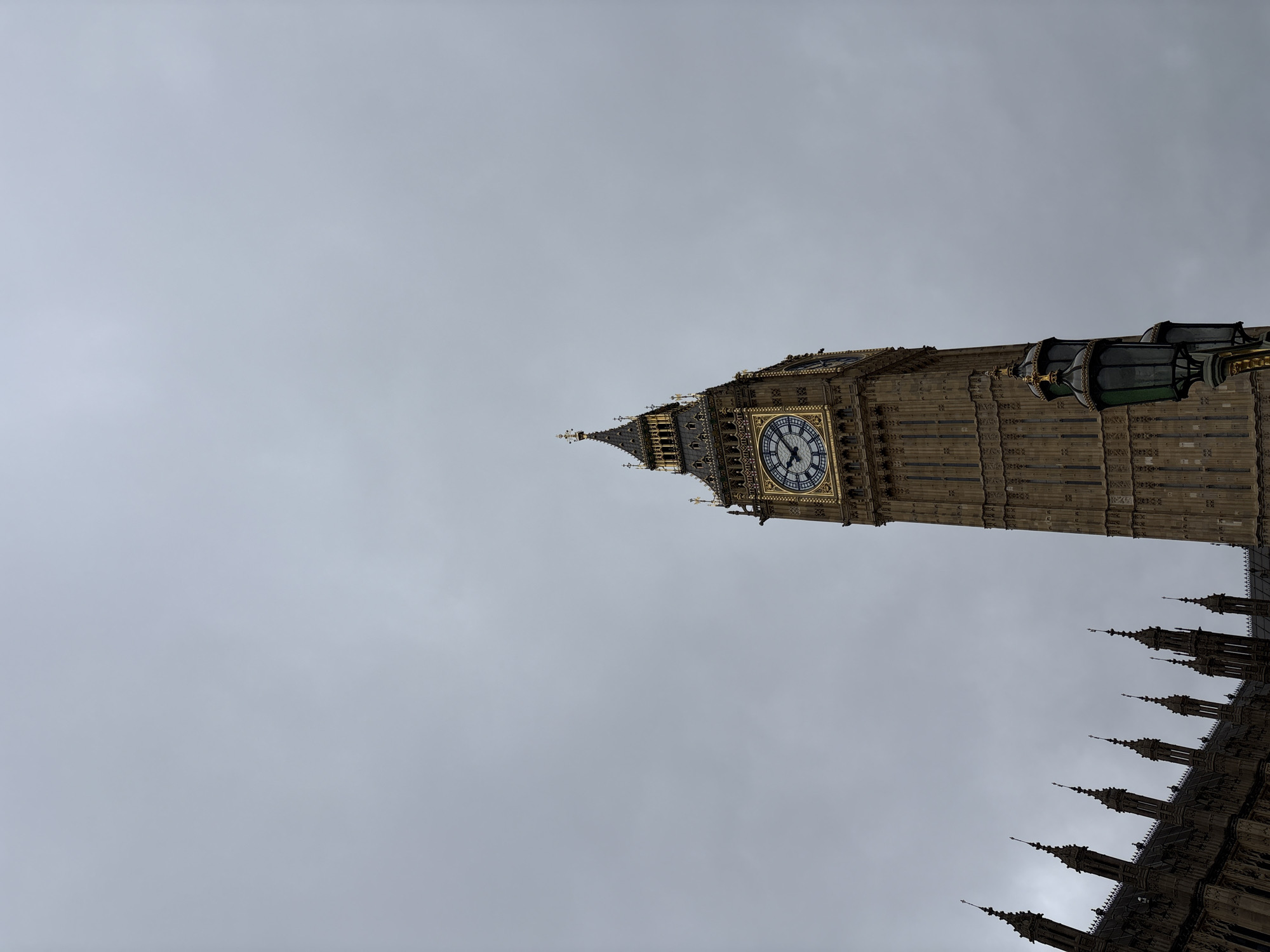 The image depicts a low-angle view of the Elizabeth Tower, commonly known as Big Ben, in London, United Kingdom, set against an overcast sky. The iconic clock tower is prominently featured, showing its intricate architectural details and the clock face. Parts of the Palace of Westminster's roof are visible on the right side. There are no people in the shot.