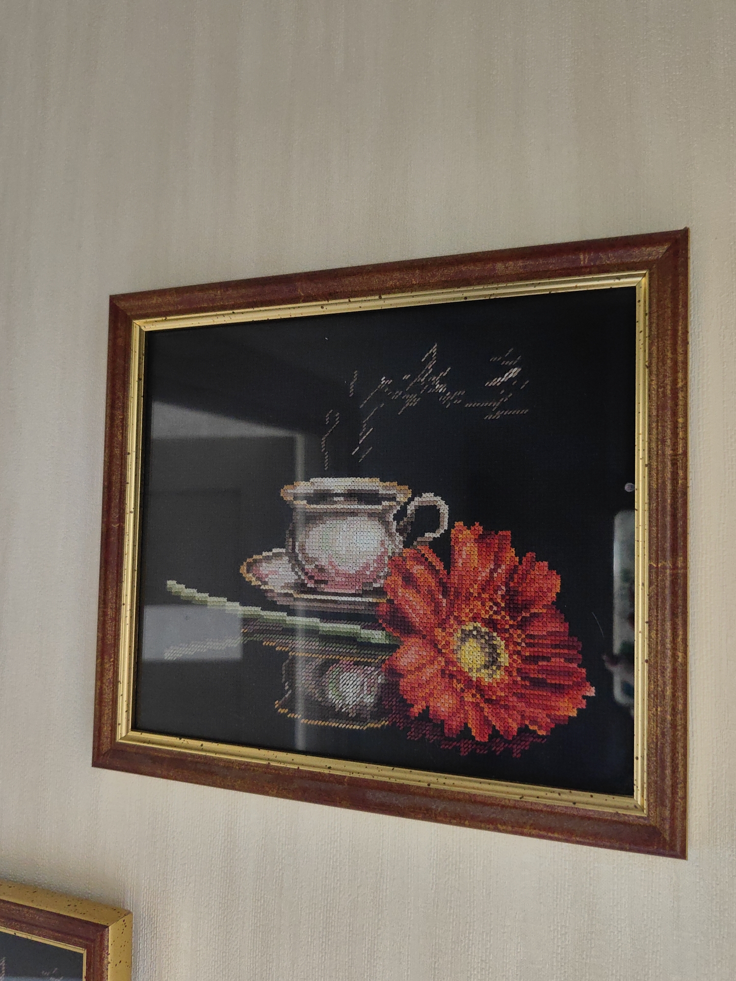 The image features a framed cross-stitch artwork hanging on a textured, light beige or off-white wall. The artwork itself is a still life set against a dark background, depicting a white teacup or coffee cup on a saucer, with faint steam rising from it. To the right of the cup, a large, vibrant red-orange flower, possibly a gerbera daisy, with a prominent yellow center, dominates the foreground. Above the cup, delicate light-colored cursive script is stitched into the artwork.
The cross-stitch is protected by glass, which exhibits reflections from the surrounding environment, particularly noticeable on the left side of the frame. The frame itself is rectangular, with a reddish-brown outer edge and a narrower golden inner trim, appearing slightly aged or distressed. The wall has a subtle, vertical pattern, suggesting wallpaper. In the lower-left corner of the photograph, a small portion of another framed item, with a golden frame, is visible below the main artwork. There are no people visible, and no specific activity or event is taking place beyond the display of the artwork. Time of day, weather, and emotions are not discernible from the image. No explicit location cues for Sochi, Russia are visible within the frame.