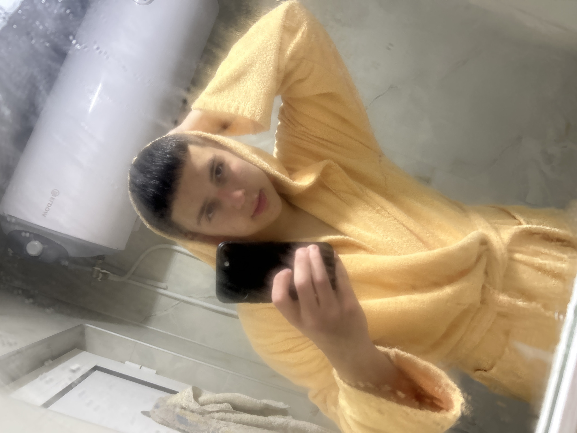 A mirror selfie taken in Zlatograd, Bulgaria, features a young person, likely male, wearing a soft yellow hooded bathrobe. The individual holds a black smartphone in their right hand, capturing their reflection, while their left arm is raised casually behind their head. They have short, dark hair and a calm, neutral expression.
The setting appears to be a functional domestic space, possibly a bathroom or utility room. Reflected prominently behind the person is a large, white, cylindrical water heater or boiler, positioned towards the left of the frame. The brand name "ERDOW" is clearly visible, inverted, near the base of the heater. White pipes or conduits run horizontally across the background walls. To the lower left, a rectangular, light-colored structure, possibly a window frame or part of a fixture, is visible. A light-colored towel or cloth is also seen near the bottom of the reflection. The surfaces in the background are light-toned, suggesting tiled walls or a similar finish. The bright lighting suggests the image was taken during daytime.