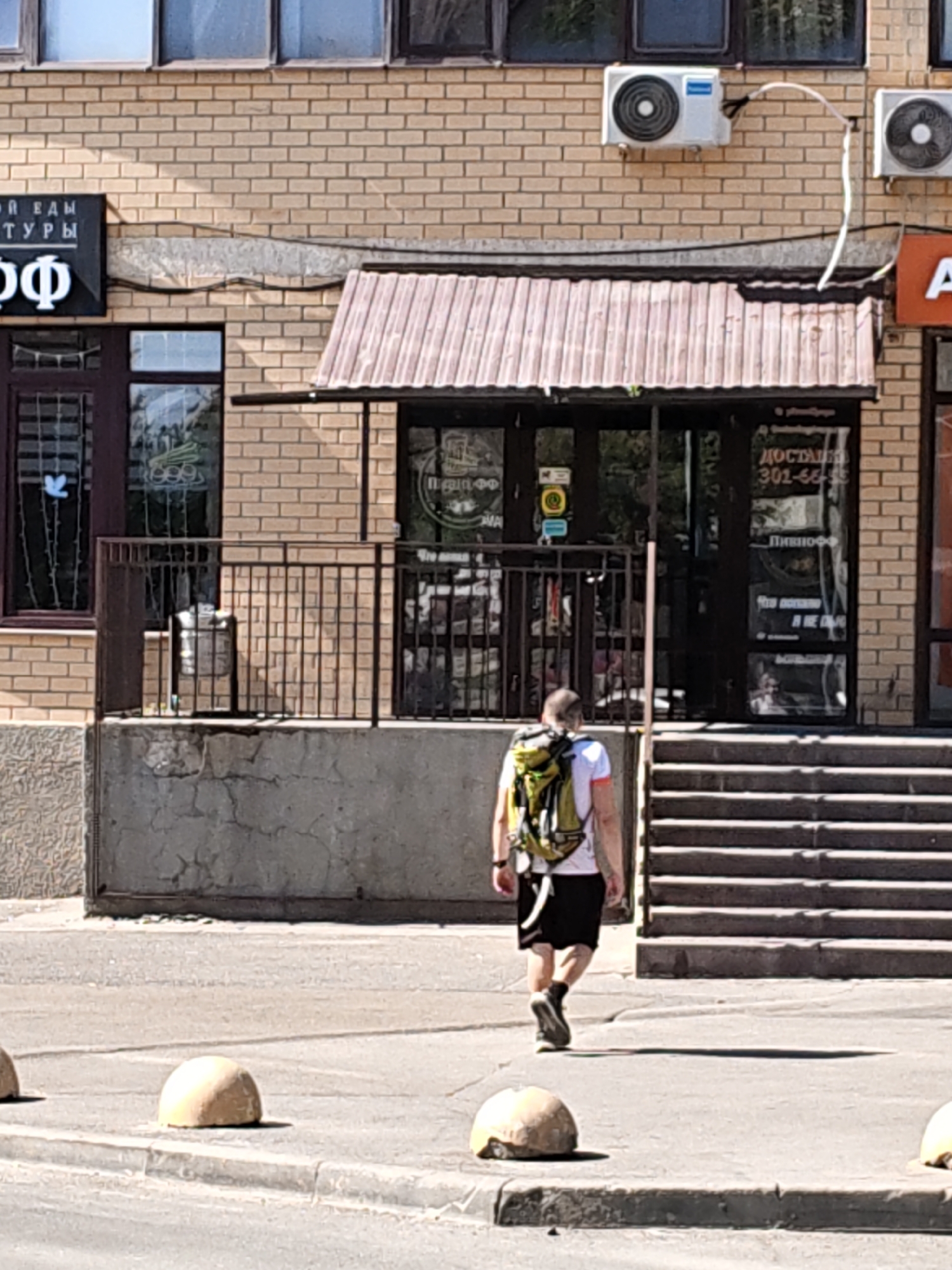 A man with a yellow and green backpack walks away from the camera, towards a building with a red-tiled awning. He is wearing a white t-shirt with orange trim and black shorts. The building has a brick facade and several windows. On the left side of the building, there is a black sign with white text that reads "КАМ ТУРЫ" and "Ф". On the right side of the awning, there are two air conditioning units mounted on the brick wall. Below the awning, there is a glass door with signs on it. To the right of the door are a set of concrete stairs. In the foreground, there are three concrete bollards in a row on the side of a road. The scene appears to be on a sunny day. This image was taken in Rostov-on-Don, Russia.