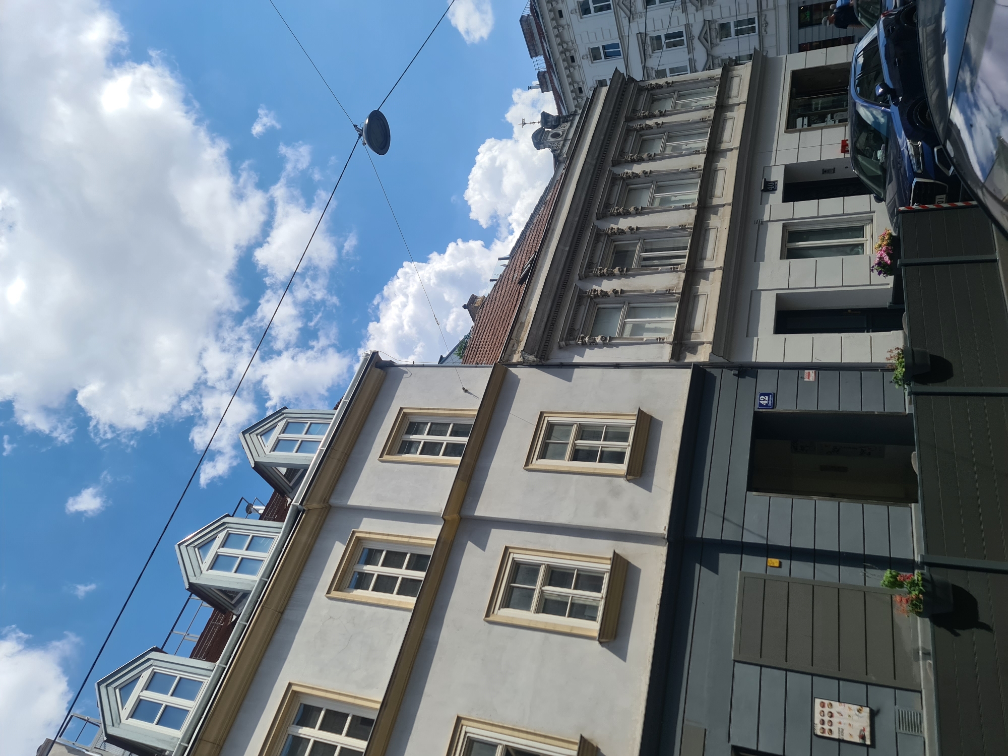 This is a street-level, vertical, full-shot photograph of buildings in Vienna, Austria, taken on a sunny day with scattered clouds. The focus is on the facades of two adjacent buildings. The building on the left is lighter in color with several windows. The building on the right has a more ornate facade with multiple windows. Above the buildings, electrical wires and a streetlamp are visible against the blue sky.
In the foreground, there is a dark gray, modern-looking facade with a prominent horizontal design. Two recessed entrances are visible, one to the left and one to the right, revealing dark interiors. A street sign with the number "42" and Cyrillic text is affixed to the right building. Planters with flowers are positioned in front of the gray facade. A dark blue SUV is parked in front of the building on the right, partially obscuring another vehicle. A few people are visible on the sidewalk in the distance.
The scene is composed of architectural elements typical of a European city, with a contrast between older, ornate buildings and more modern street-level features. The lighting suggests it is midday.