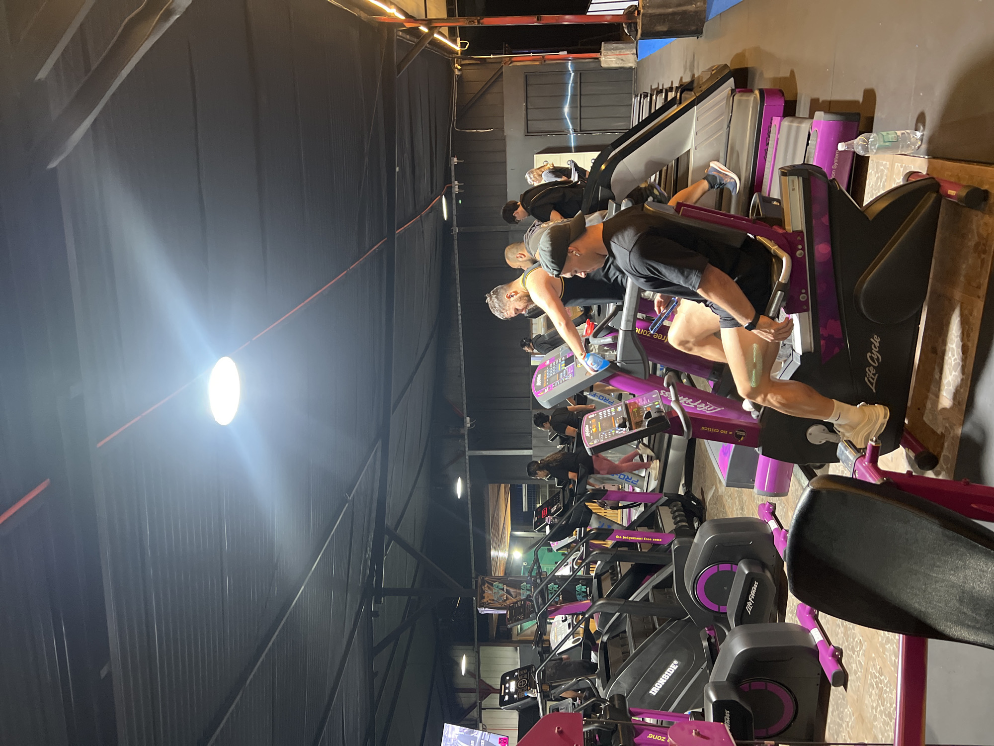This indoor shot captures a row of exercise machines, including treadmills and elliptical trainers, in what appears to be a gym or fitness center. Several individuals are engaged in exercising on the machines. The equipment is primarily black and purple, with the Life Fitness logo visible on some machines. Lighting fixtures are visible on the ceiling. While the city of Arica, Chile, is mentioned in the query, there is no direct information in the image to corroborate its location.