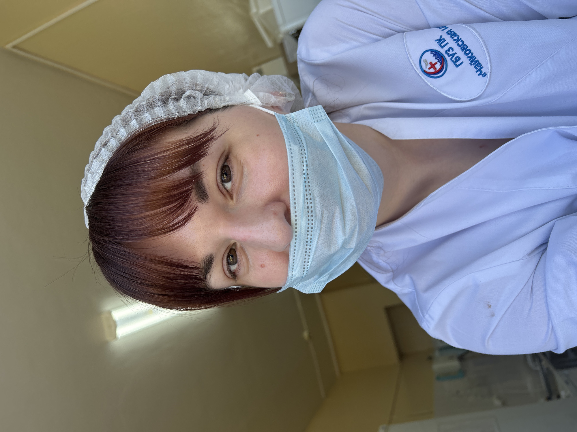 The image shows a woman in a medical setting, likely in Фоки, Russia based on the Cyrillic text on her white coat. She is wearing a white surgical cap and a light blue surgical mask that covers her mouth and nose. Her hair is reddish-brown with bangs. She is wearing a white lab coat with a patch on the upper left shoulder that includes the Cyrillic text "ГБУЗ ПК "Чайковская ЦГБ". There are some small stains on her coat. The background appears to be an interior room with plain walls and fluorescent lighting.