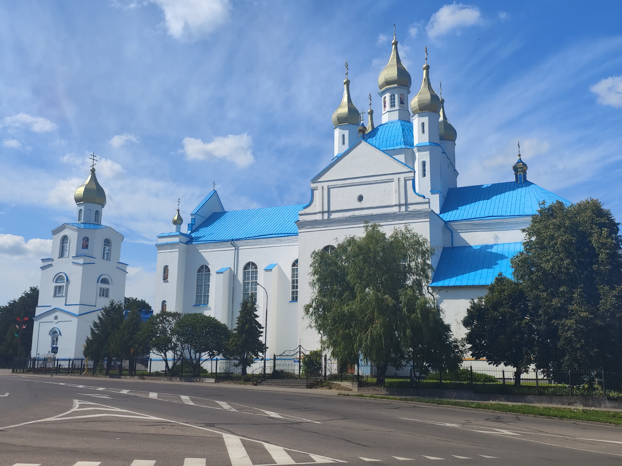 This is a daytime, eye-level, wide-angle outdoor photograph of the Saint Casimir Church in Slonim, Belarus. The church, constructed of white plaster with bright blue metal roofing, features multiple golden domes topped with crosses. The main structure is flanked by a taller bell tower on the left, also adorned with a golden dome and cross. A black metal fence runs along the property line, separating it from the paved road in the foreground. Trees, some deciduous and some coniferous, are visible on the church grounds. The sky is a vibrant blue with scattered white clouds. The road in the foreground is marked with white pedestrian crossing lines. No people are visible in the image.